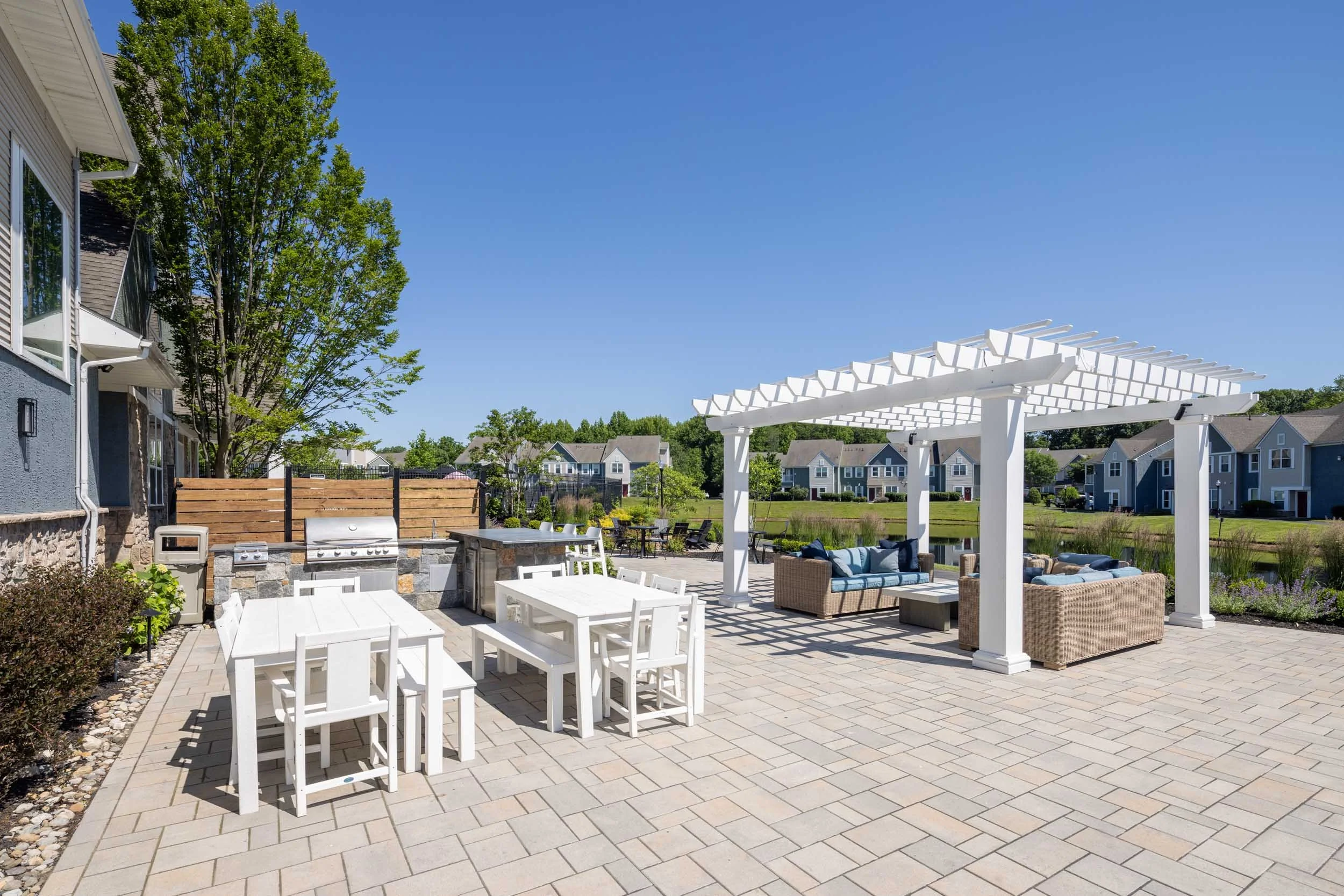 A spacious patio area with outdoor furniture, including a dining table with white chairs, a seating area with wicker sofas and cushions, and a white pergola. There is a grill and a small fire pit nearby, and surrounding the area is lush greenery and 