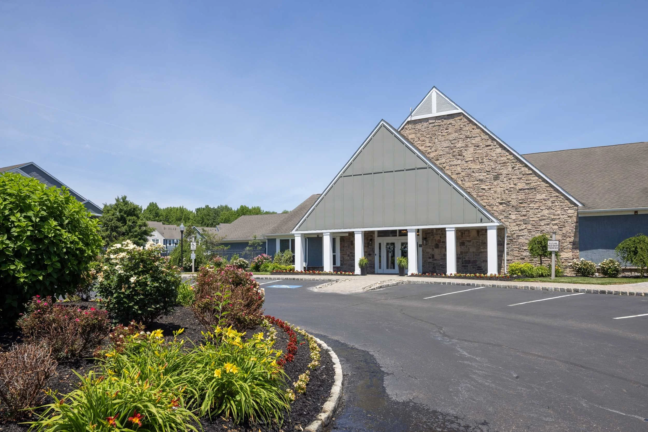 A building with a peaked roof and exterior walls made of stone and metal siding, set in a parking lot with landscaped flower beds and greenery, under a clear blue sky.