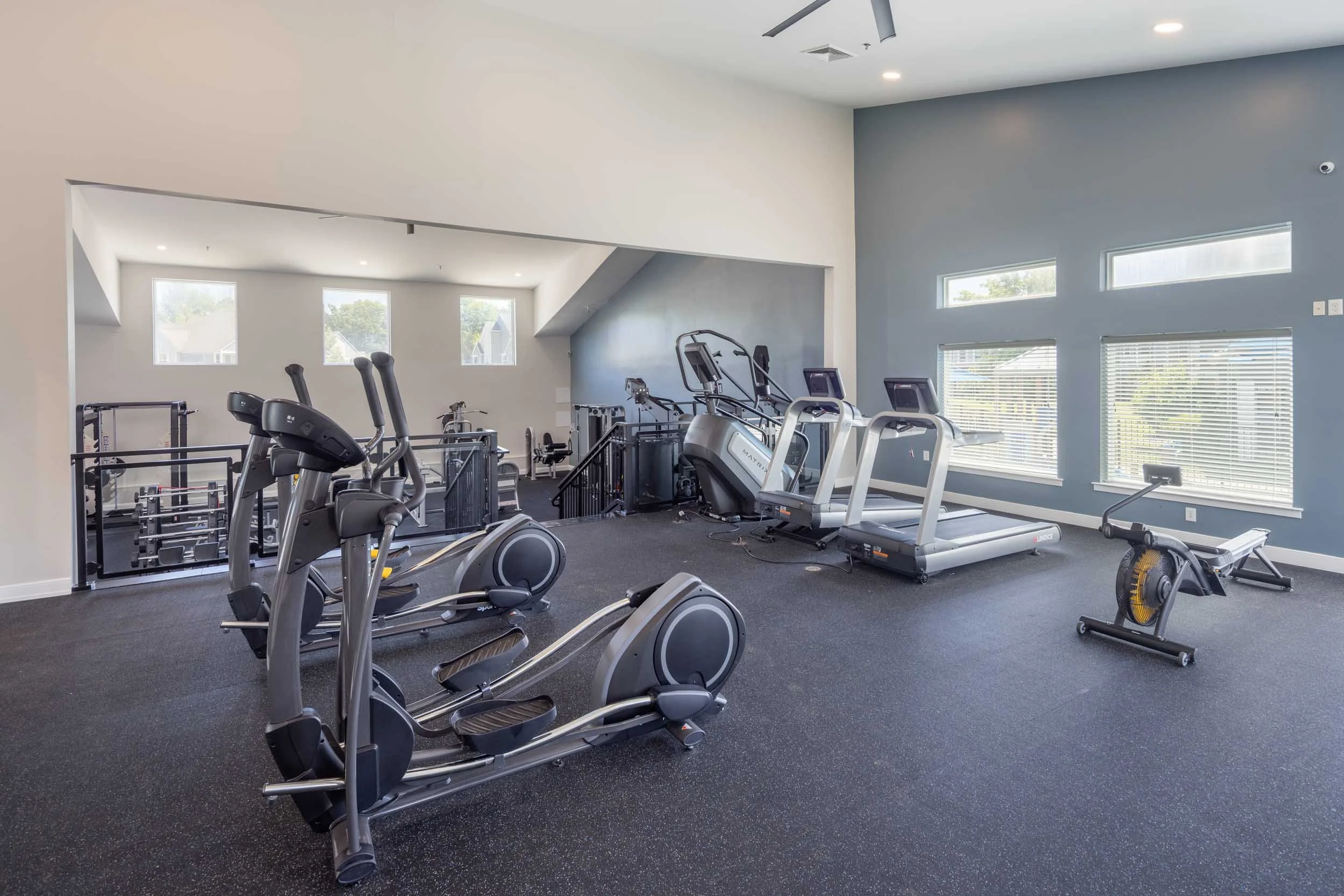 A spacious gym room with windows letting in natural light, featuring cardio and strength training equipment including treadmills, ellipticals, and weight machines.