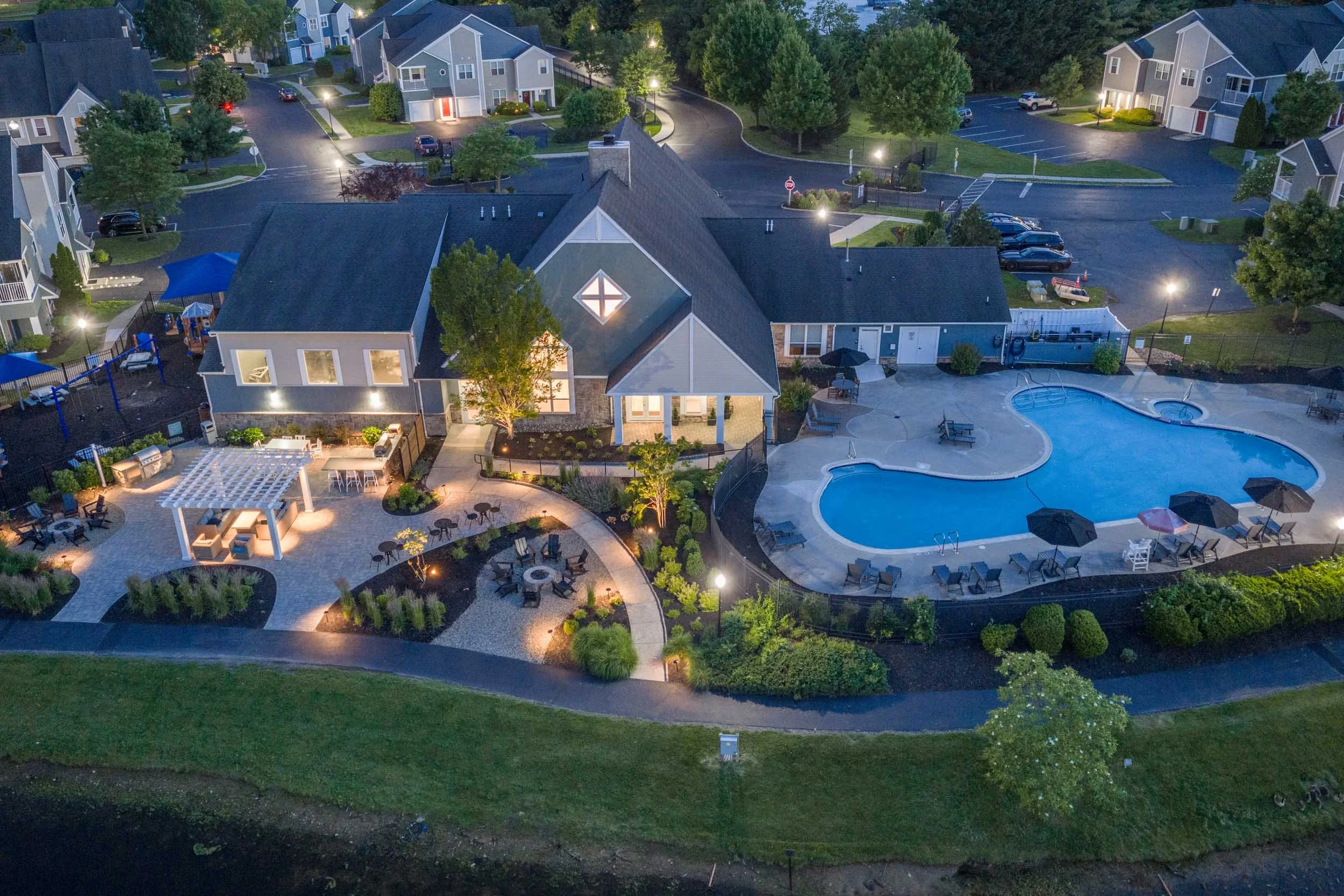 Aerial view of a residential community at dusk with houses, trees, a parking lot, and a swimming pool area illuminated by outdoor lights.