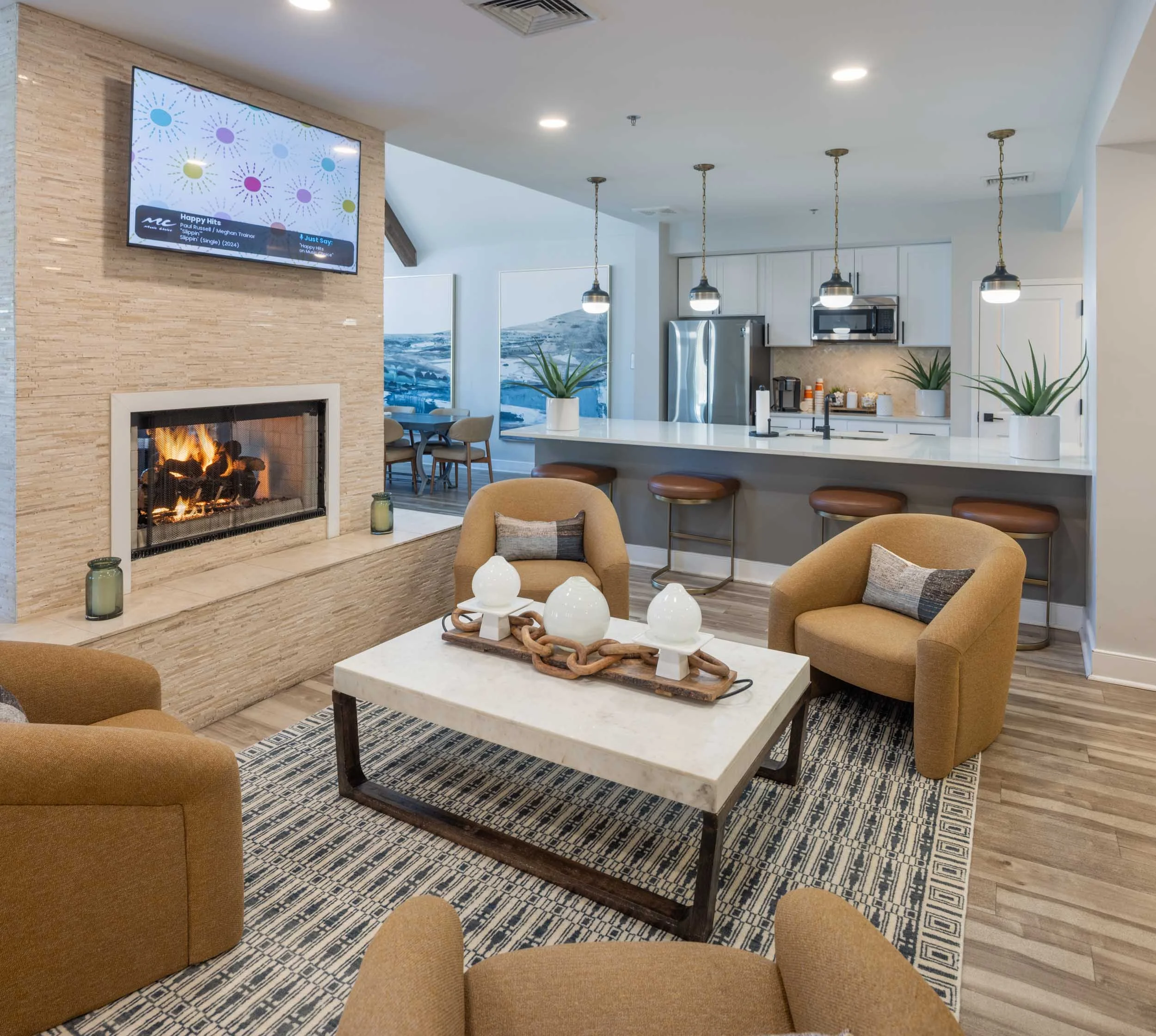 Modern kitchen and lounge with beige chairs, fireplace, flat-screen TV, kitchen island with bar stools, pendant lights, and potted plants.