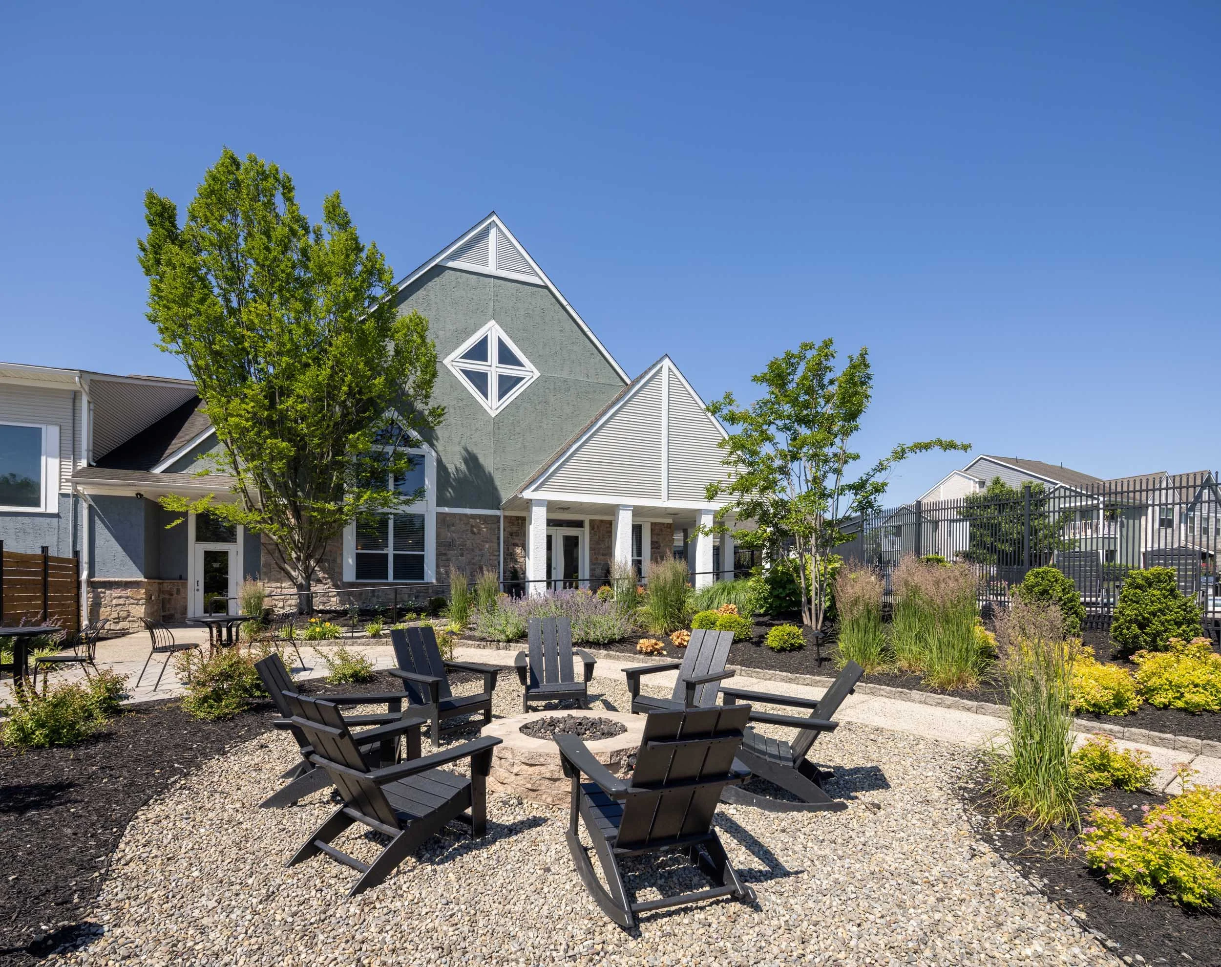A modern residential neighborhood with a landscaped backyard, featuring a fire pit surrounded by black Adirondack chairs, various green plants and trees, and houses with triangular roofs and large windows under a clear blue sky.
