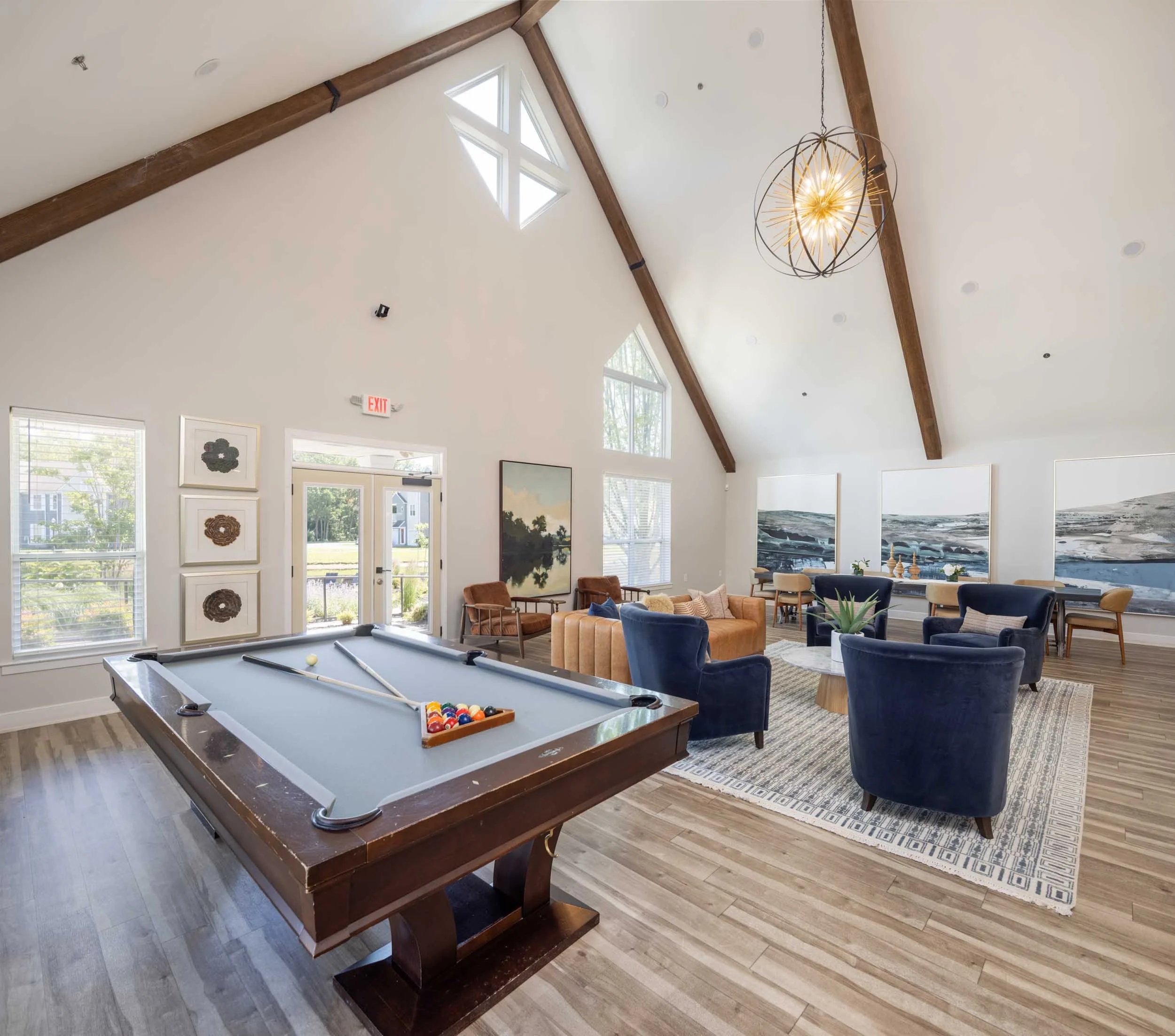Interior of a bright room with a pool table, modern chairs, and large windows, decorated with artwork and a spherical chandelier hanging from a high ceiling.