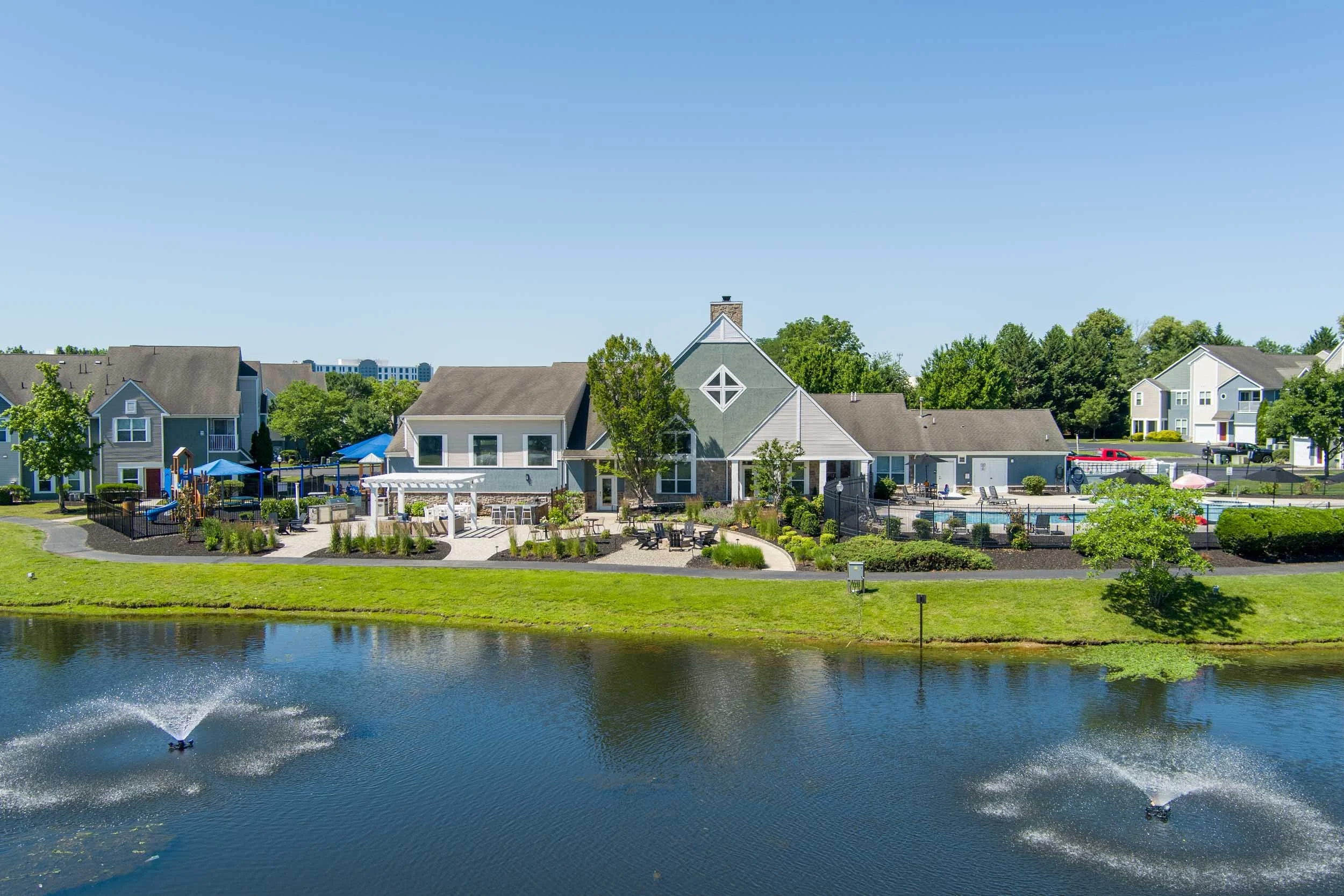 A waterfront outdoor community area with playground equipment, seating, and a swimming pool, bordered by residential houses, with a small pond or lake in the foreground and a clear blue sky above.