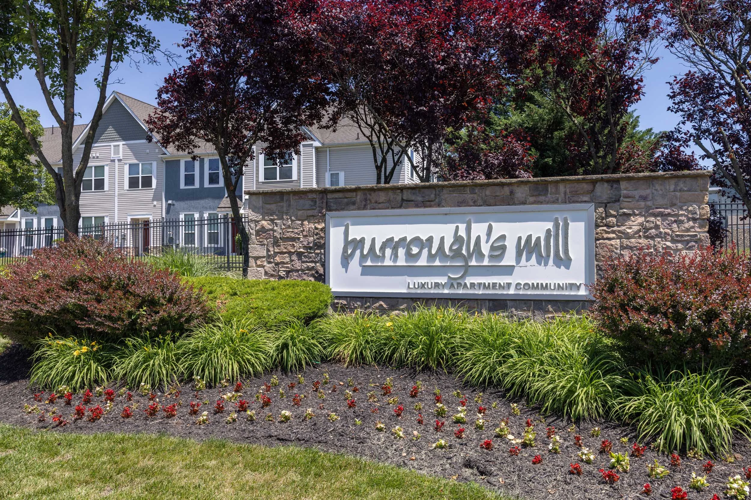 A landscaped entrance with a stone sign that reads "Burroughs Mill Luxury Apartment Community," surrounded by colorful plants and red-leaved trees, with residential buildings in the background.