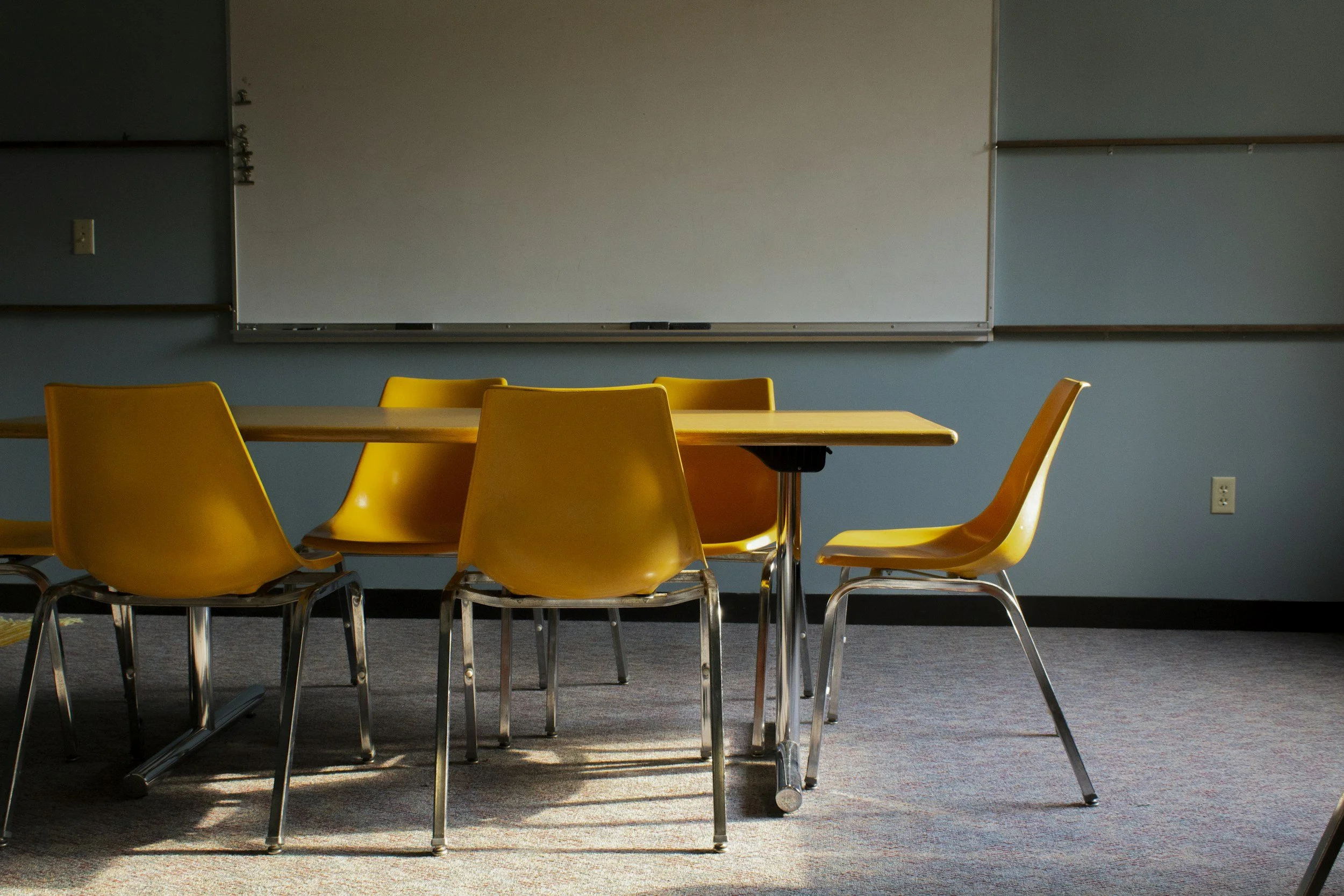 Empty classroom with yellow chairs arranged around a wooden table in front of a whiteboard on a blue wall.