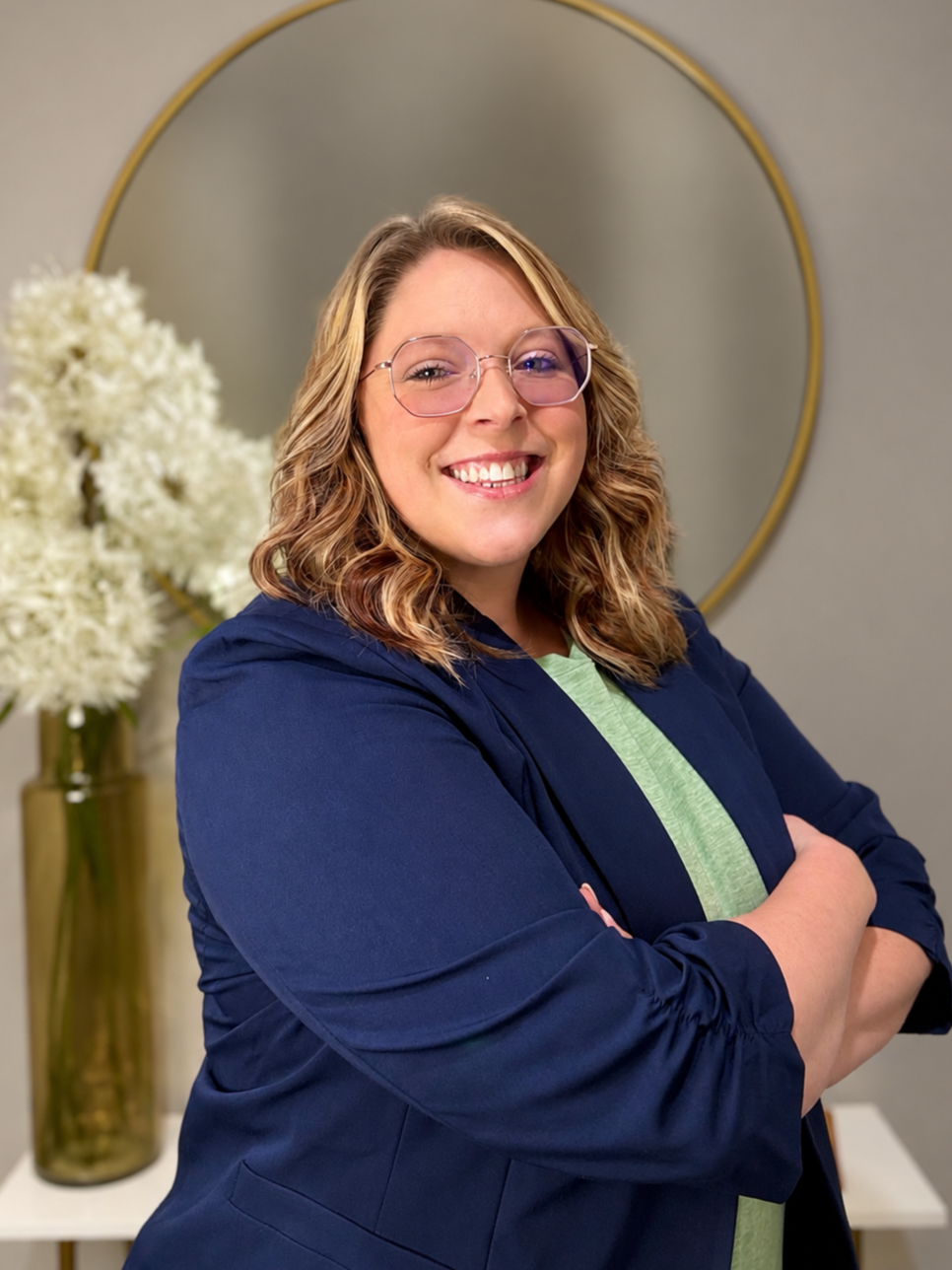 A smiling woman with curled hair, shoulder-length blonde hair and glasses, standing with arms crossed in front of a round mirror and a tall vase with dandelion flowers.
