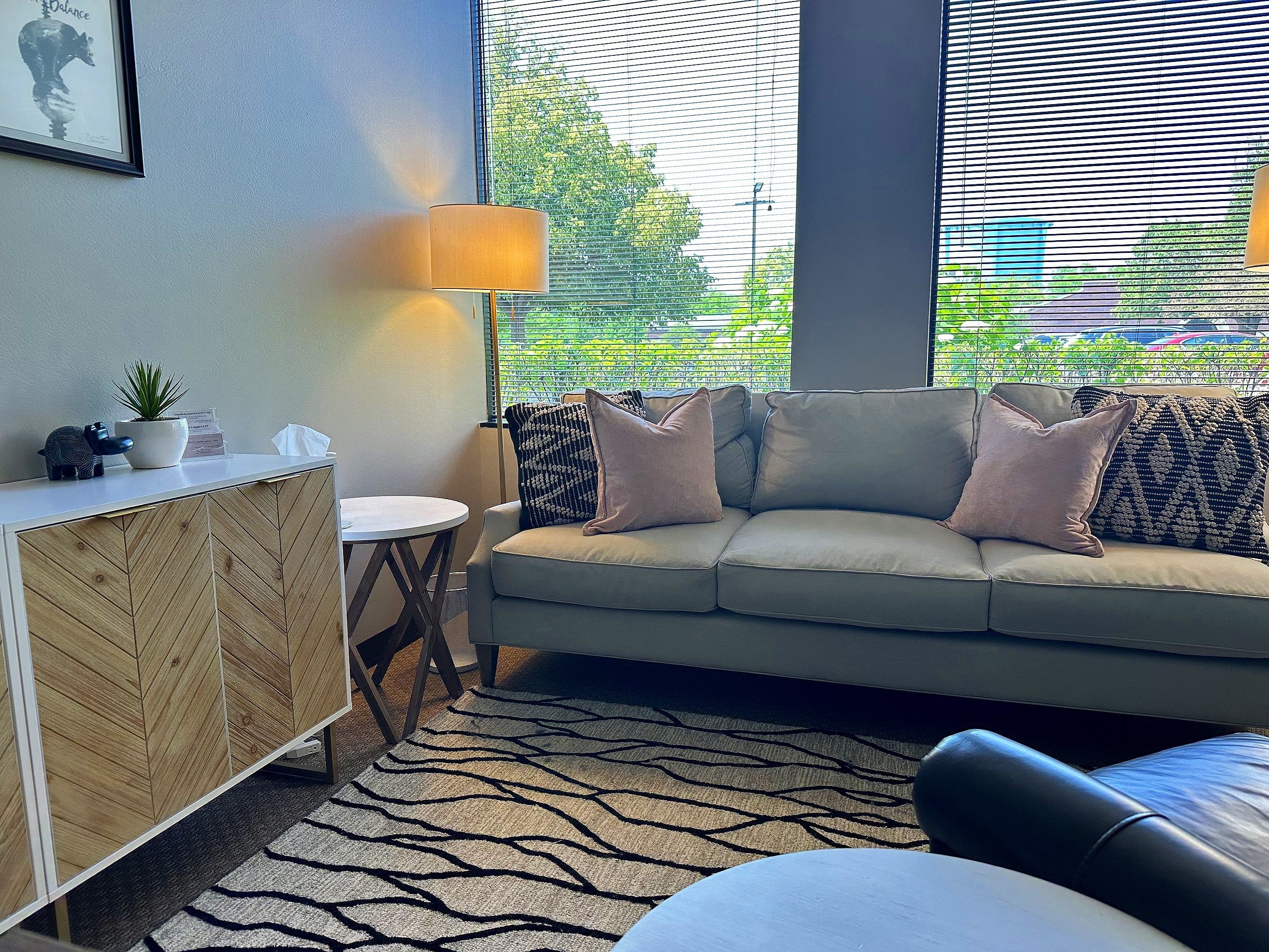 A bright living room with large windows covered with horizontal blinds, a white couch with multiple cushions, a wooden side table, a lamp, a patterned rug, and decorative items on a white cabinet.