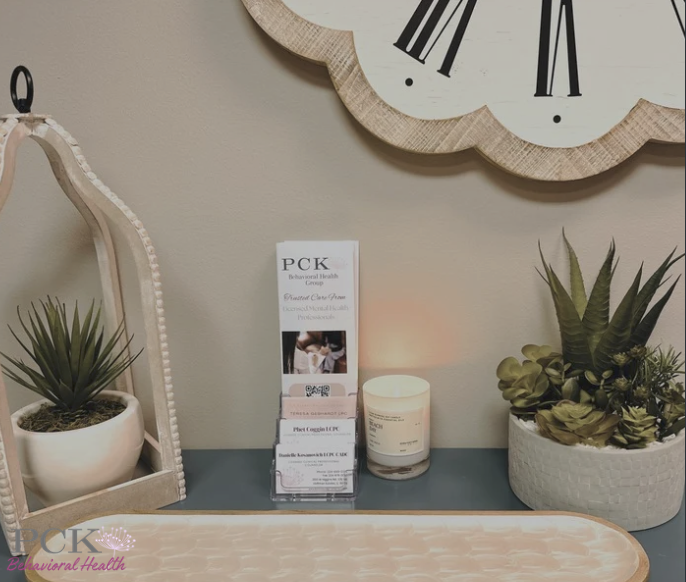 A wall clock with Roman numerals hanging above a tabletop, which has a small lamp, a brochure holder, a scented candle, and potted succulent plants.