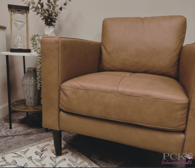 Close-up of a tan leather armchair with black wooden legs in a living room with a patterned rug, beside a small side table with a decorative vase and an hourglass, and a wall with a plant.