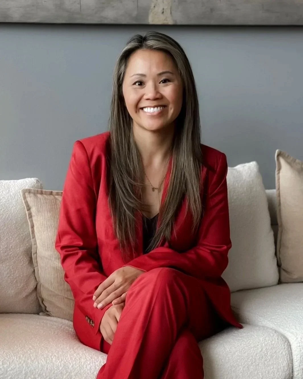 A woman with long brown hair, smiling, sitting on a sofa in a living room, wearing a red blazer and matching pants.