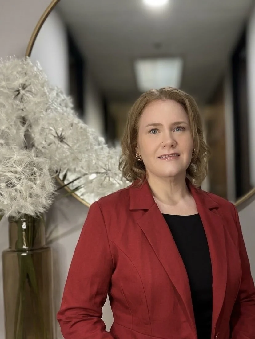 A woman with shoulder-length curly blonde hair, wearing a red blazer over a black top, standing indoors with a large mirror and a vase with white dandelion-like flowers in the background.