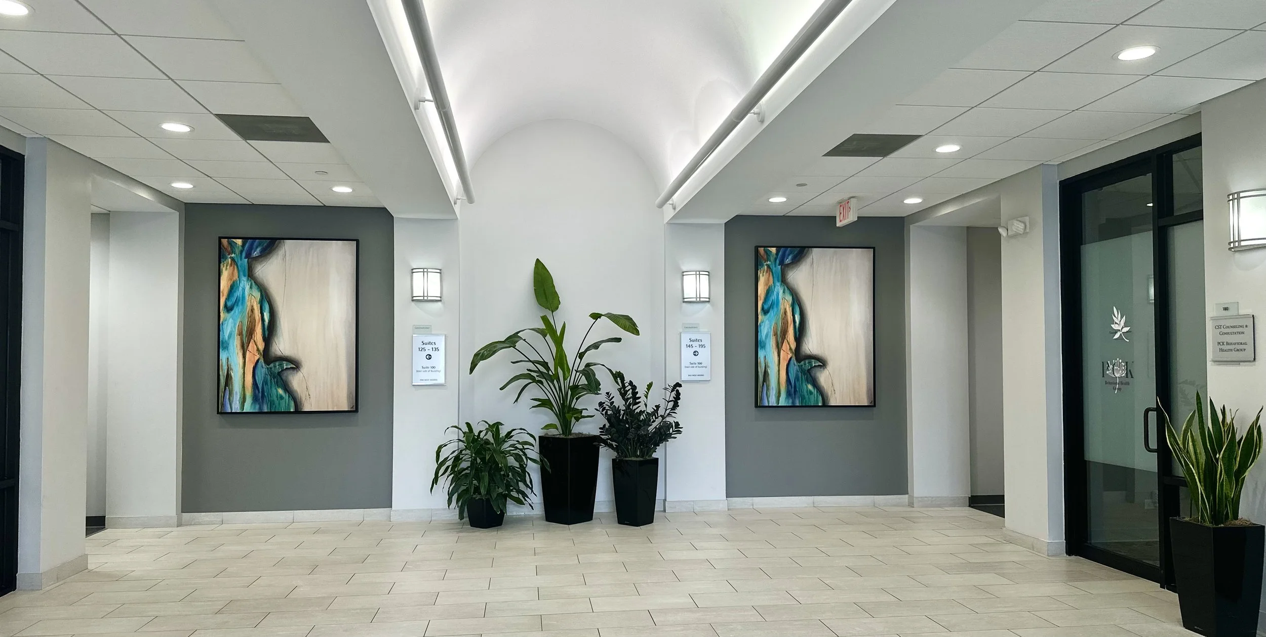 Inside a modern building with white walls, a white ceiling with recessed lighting, and tiled flooring. There are two abstract paintings on the wall, and several green potted plants in black planters. Signs indicate room numbers and office suites, and a glass door with a logo and plant graphics is visible on the right.