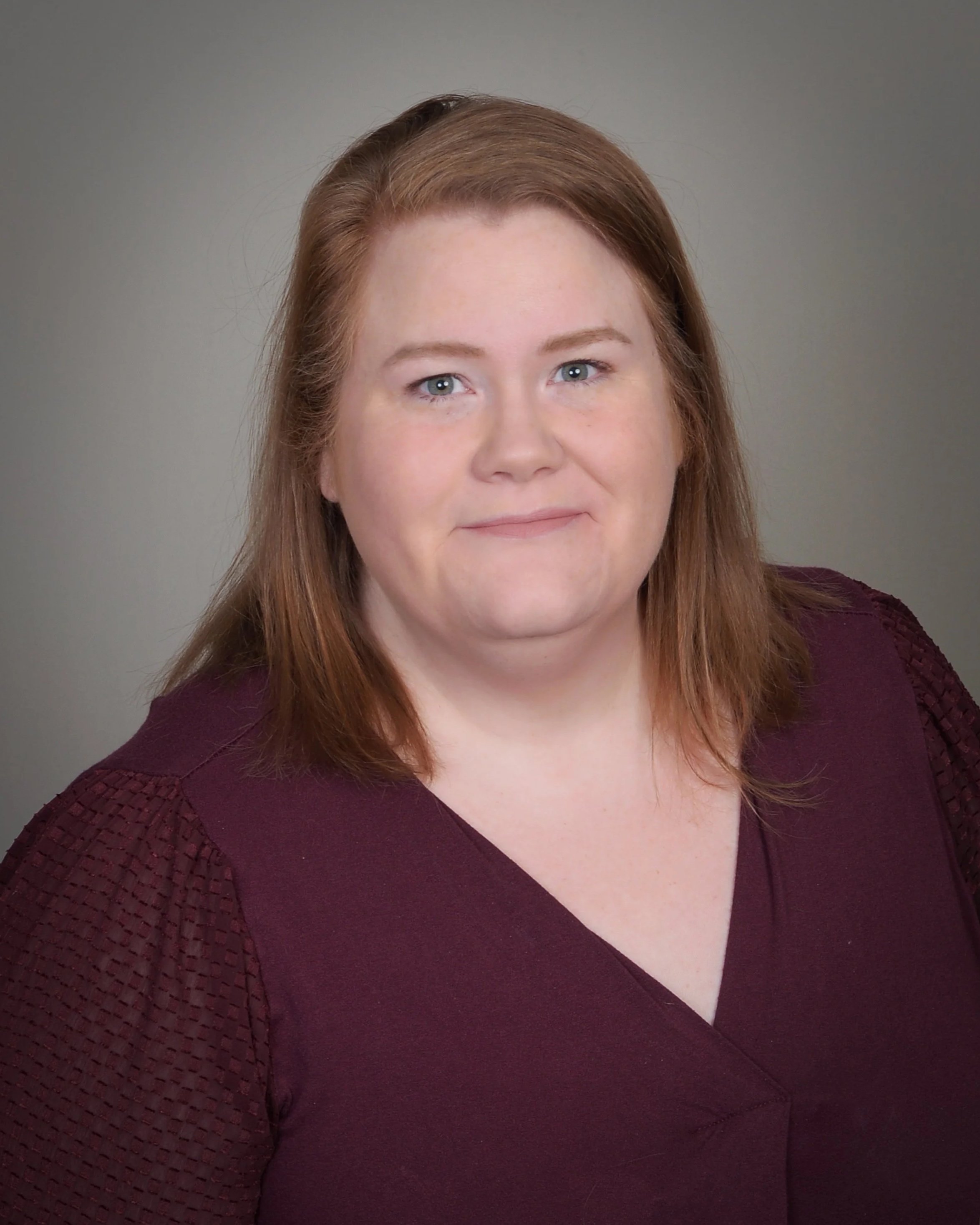 Close-up of Danielle Kosanovich with shoulder-length reddish-brown hair and blue eyes, wearing a maroon top with textured sleeves, smiling softly against a gray background.