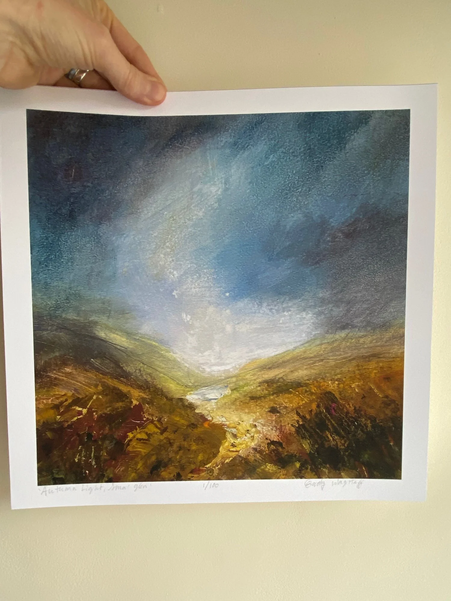 hand holding a print of light in blue sky over autumn russet glen and river