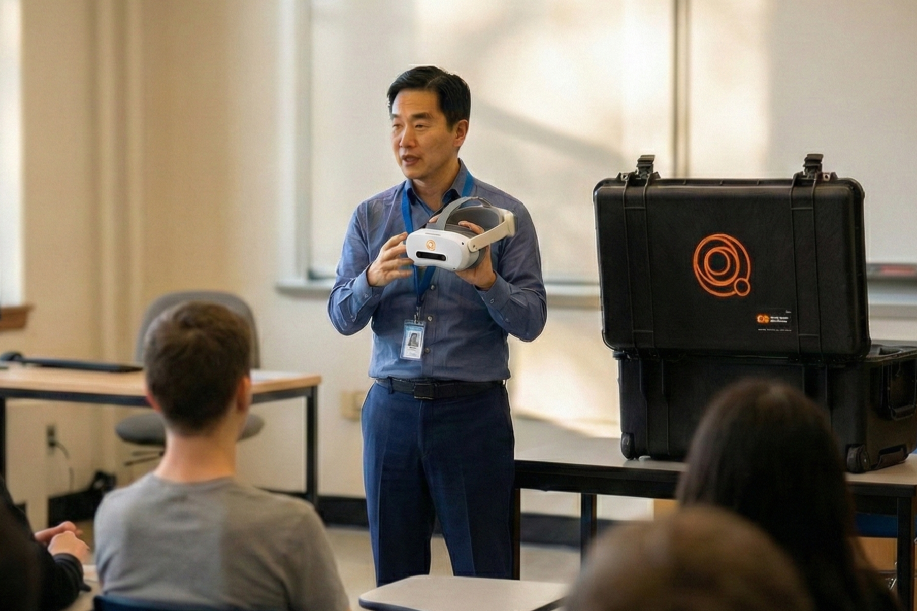 A man in a blue shirt demonstrating a virtual reality headset to an audience in a classroom setting. There is a large black case on the table next to him.