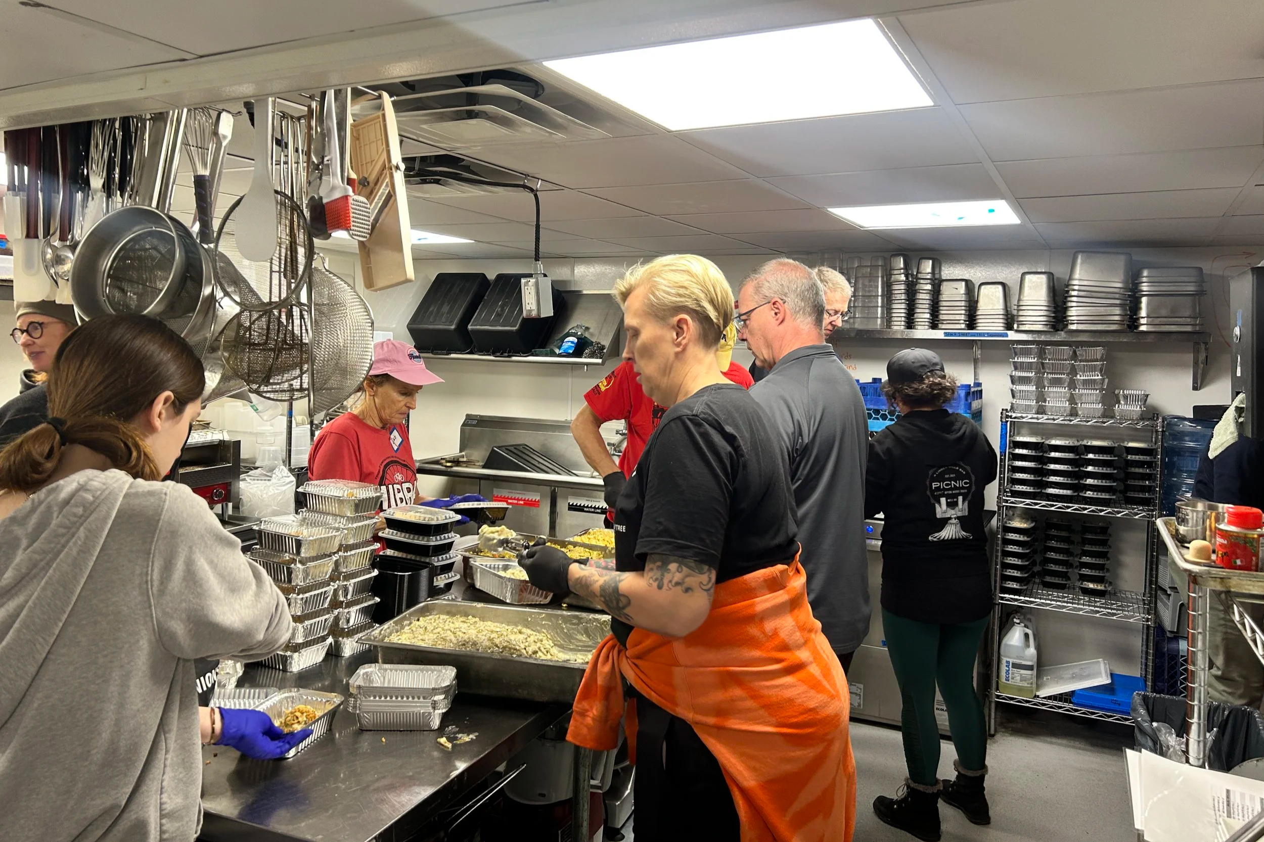 Group of people working in a commercial kitchen, preparing food at a stainless steel counter with containers and ingredients, with shelves and utensils in the background.