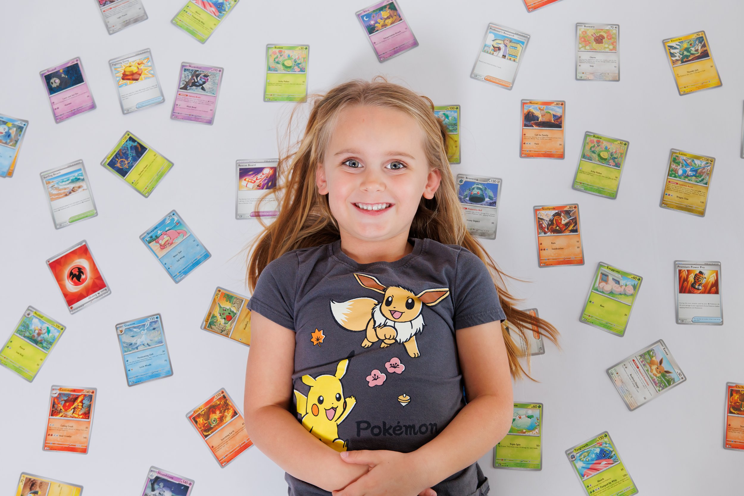 A smiling young girl with blue eyes and light brown hair lying on a white surface surrounded by numerous colorful Pokémon trading cards. She is wearing a dark gray t-shirt featuring Pokémon characters.
