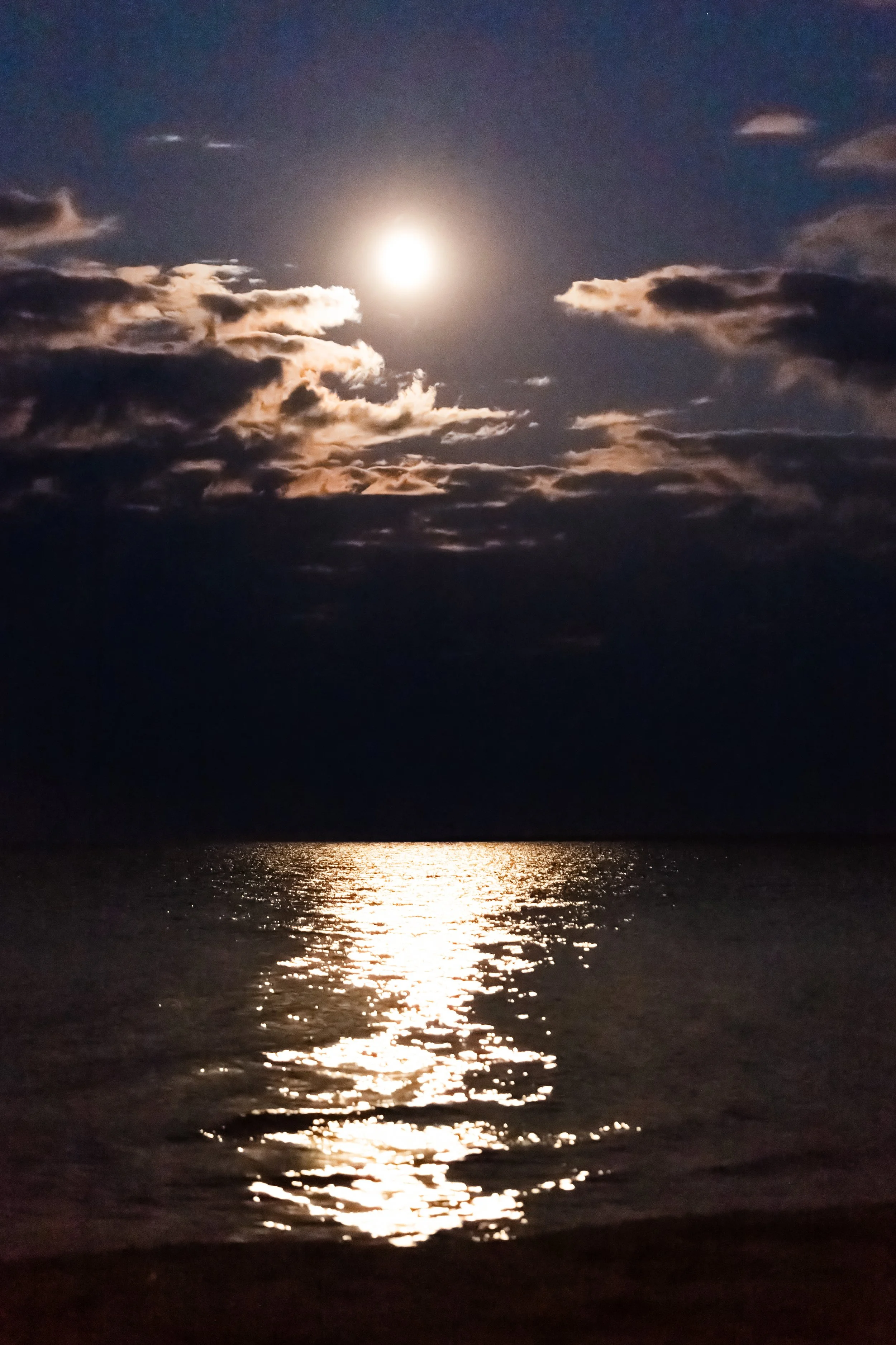 Nighttime scene of the moon reflecting on the ocean, with clouds in the sky.