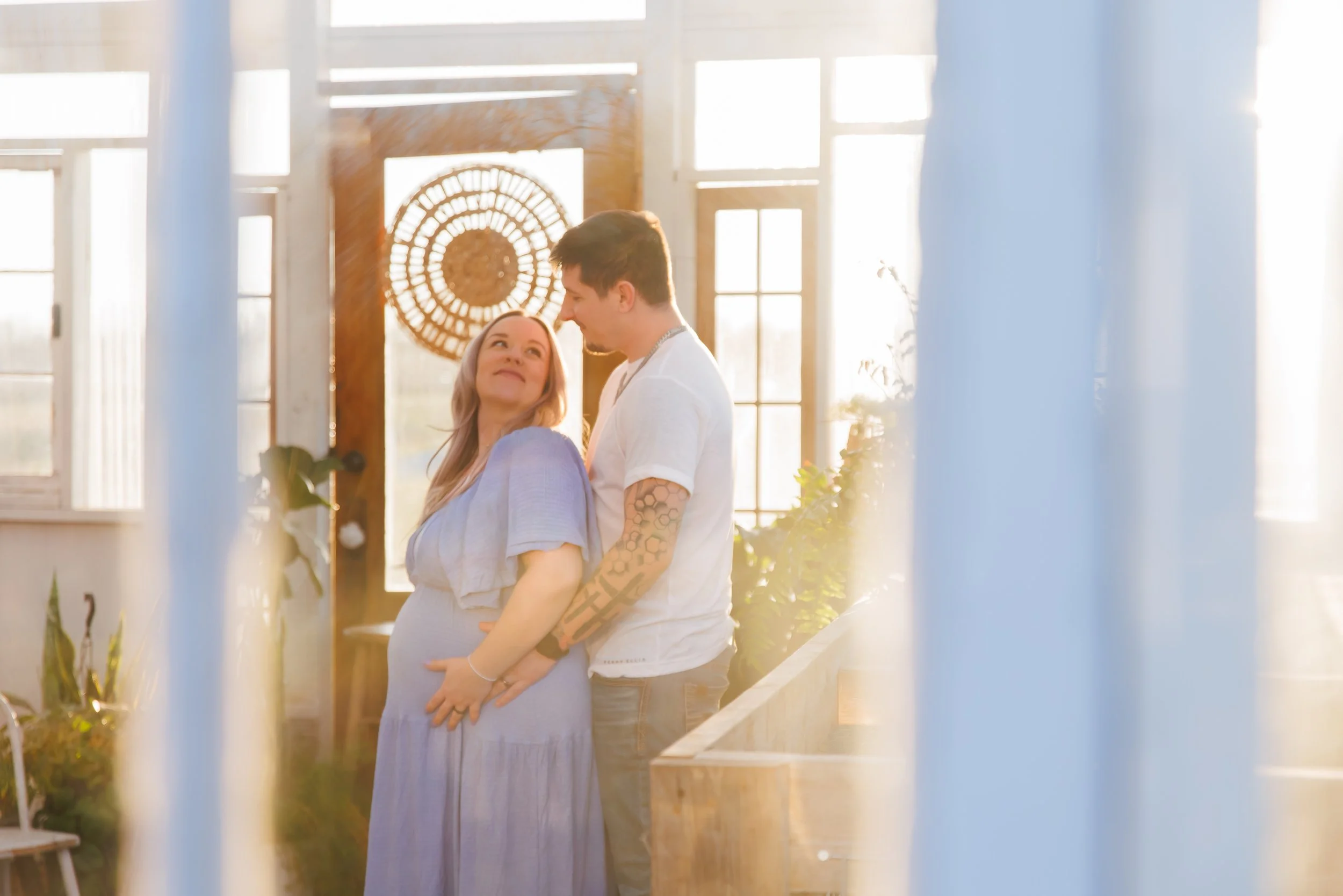 A pregnant woman and a man are standing close to each other, smiling and enjoying moment inside a sunlit room with large windows and wooden decor.