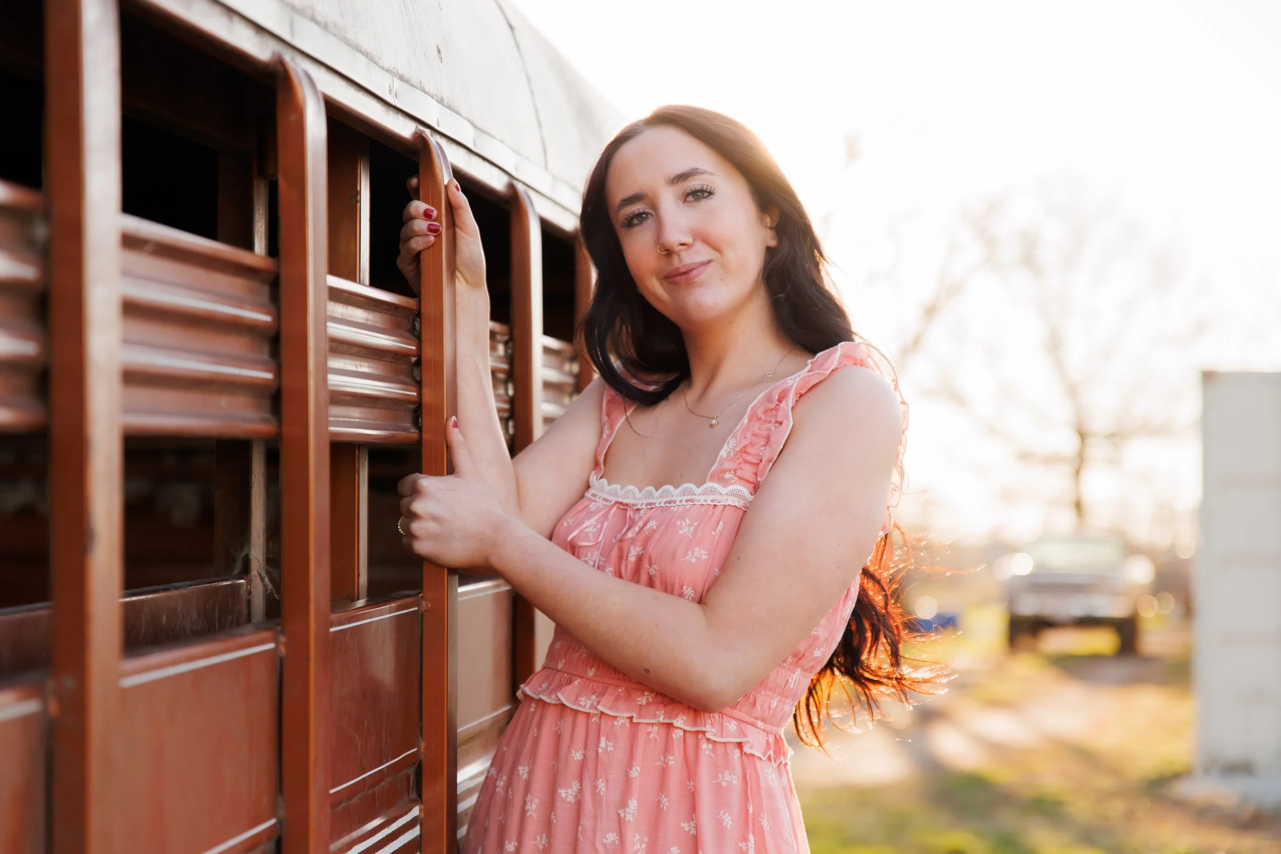 A woman with dark hair and a nose piercing wearing a pink dress with white floral patterns stands outdoors next to a wooden structure, smiling gently at the camera during golden hour with blurred background and sunlight.