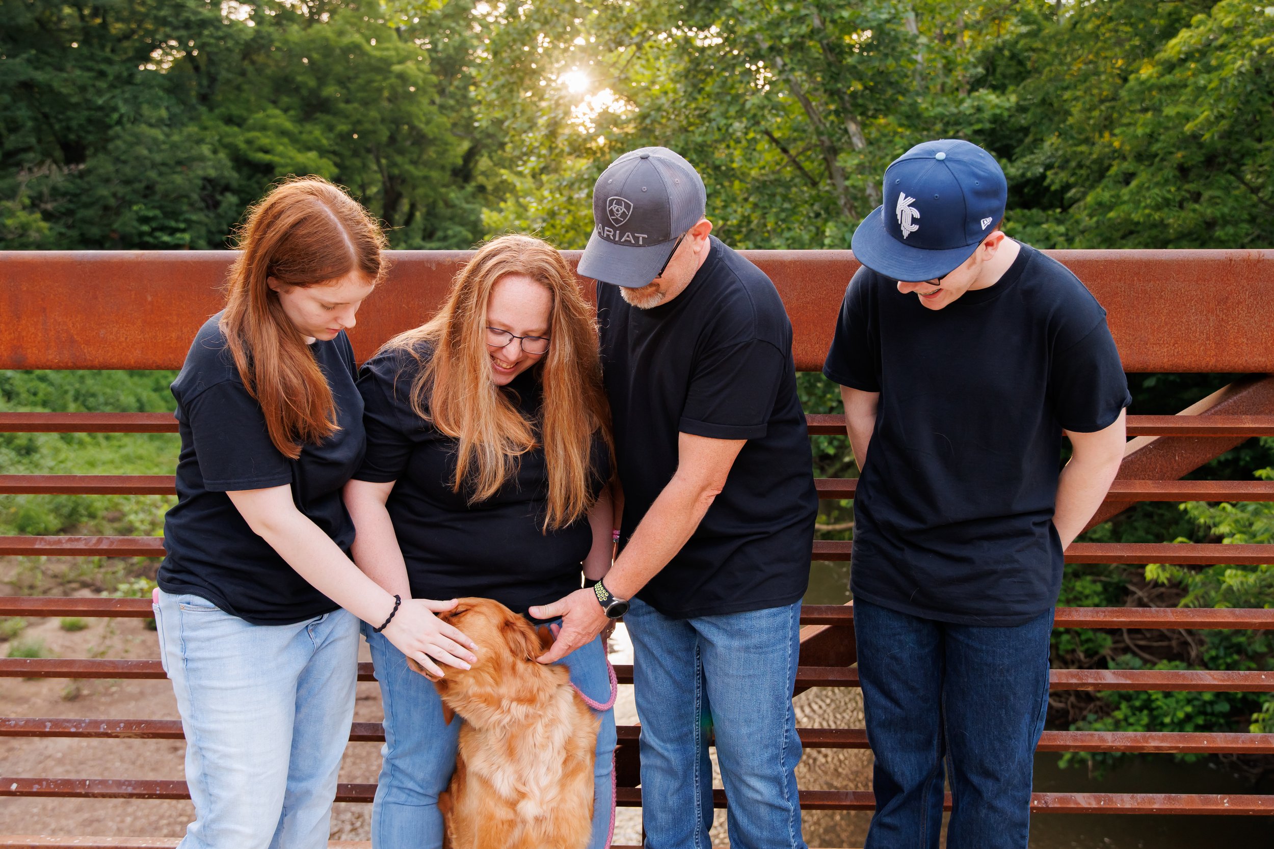 Four people, two women and two men, standing outdoors on a wooden bridge in a wooded area during sunset, petting a golden retriever dog.