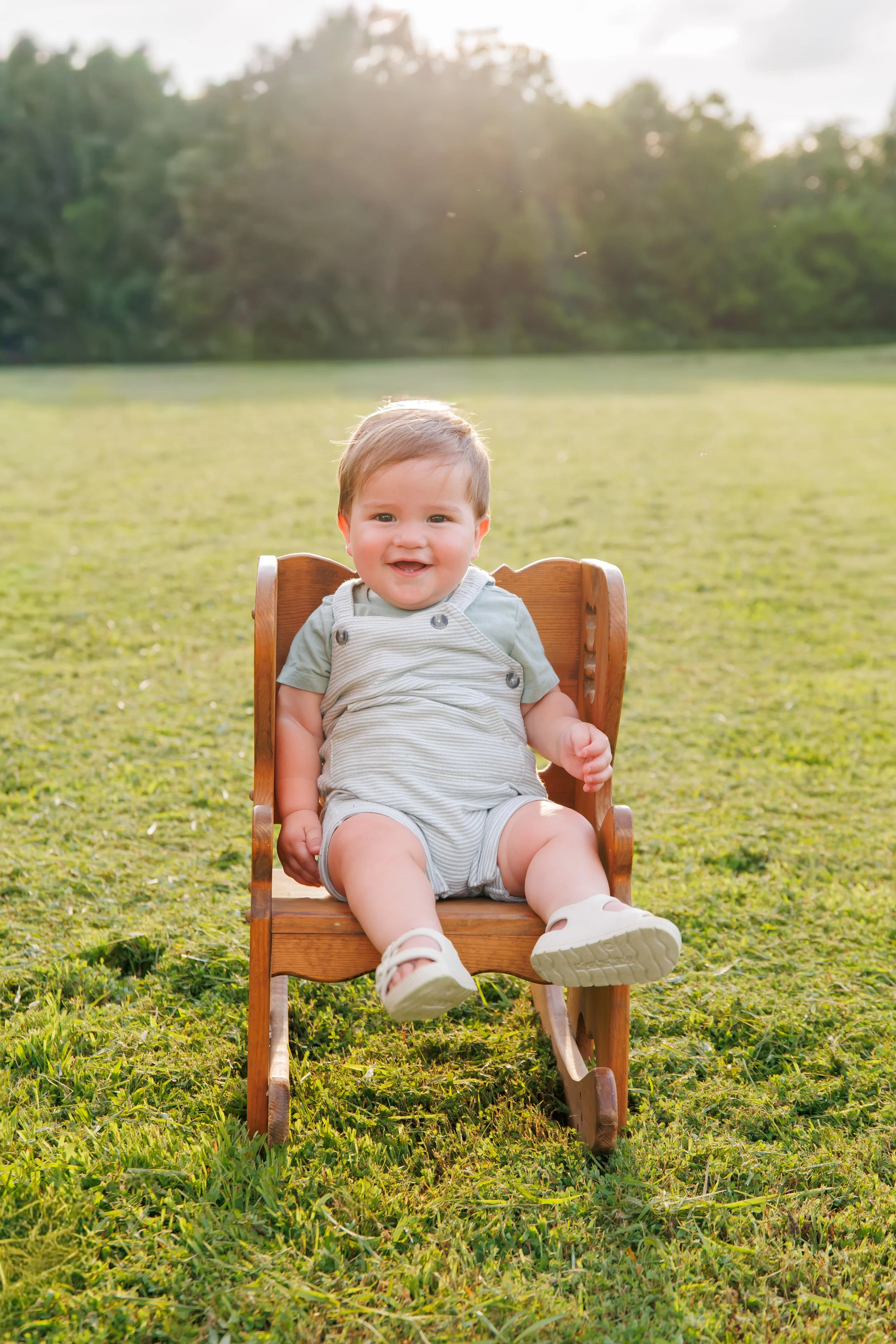 A smiling toddler sitting on a wooden lawn chair in a grassy field with trees in the background, illuminated by sunlight.