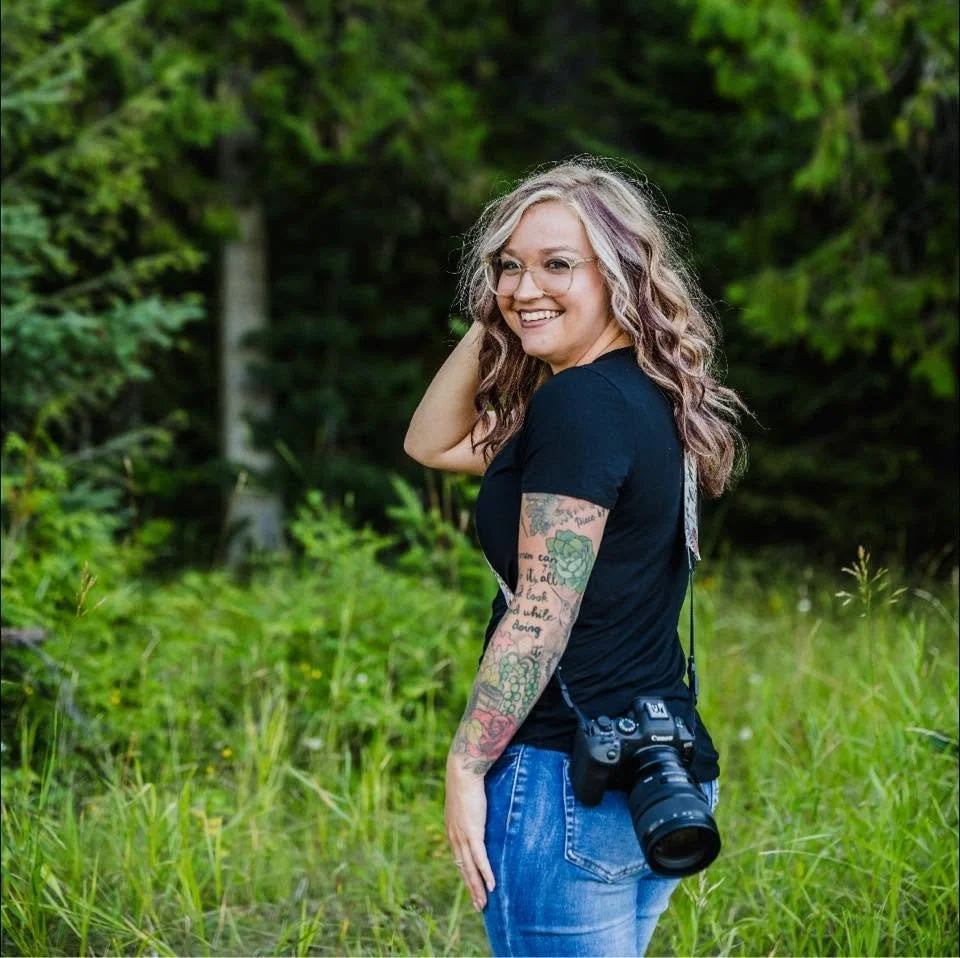 A smiling woman with curly blonde hair and tattoos on her arm, wearing glasses, a black t-shirt, and jeans, standing outdoors with a camera hanging from her neck in a lush green natural setting.