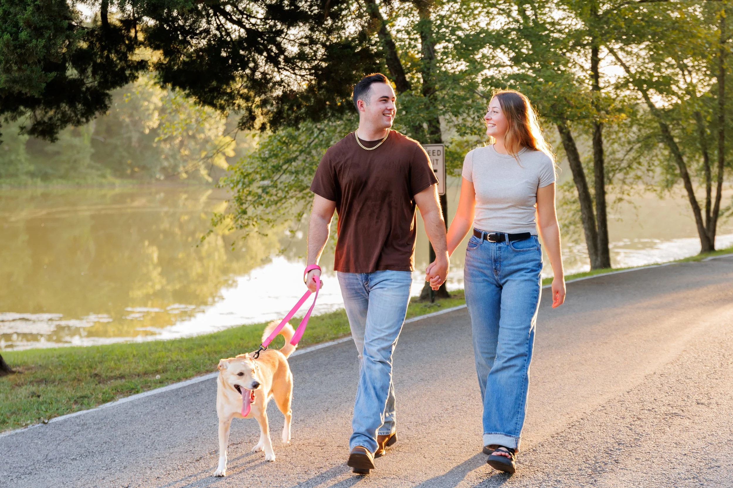 A young man and woman walking hand in hand along a park path, smiling at each other, with a service dog on a leash in front of them, during sunset by a body of water surrounded by trees.