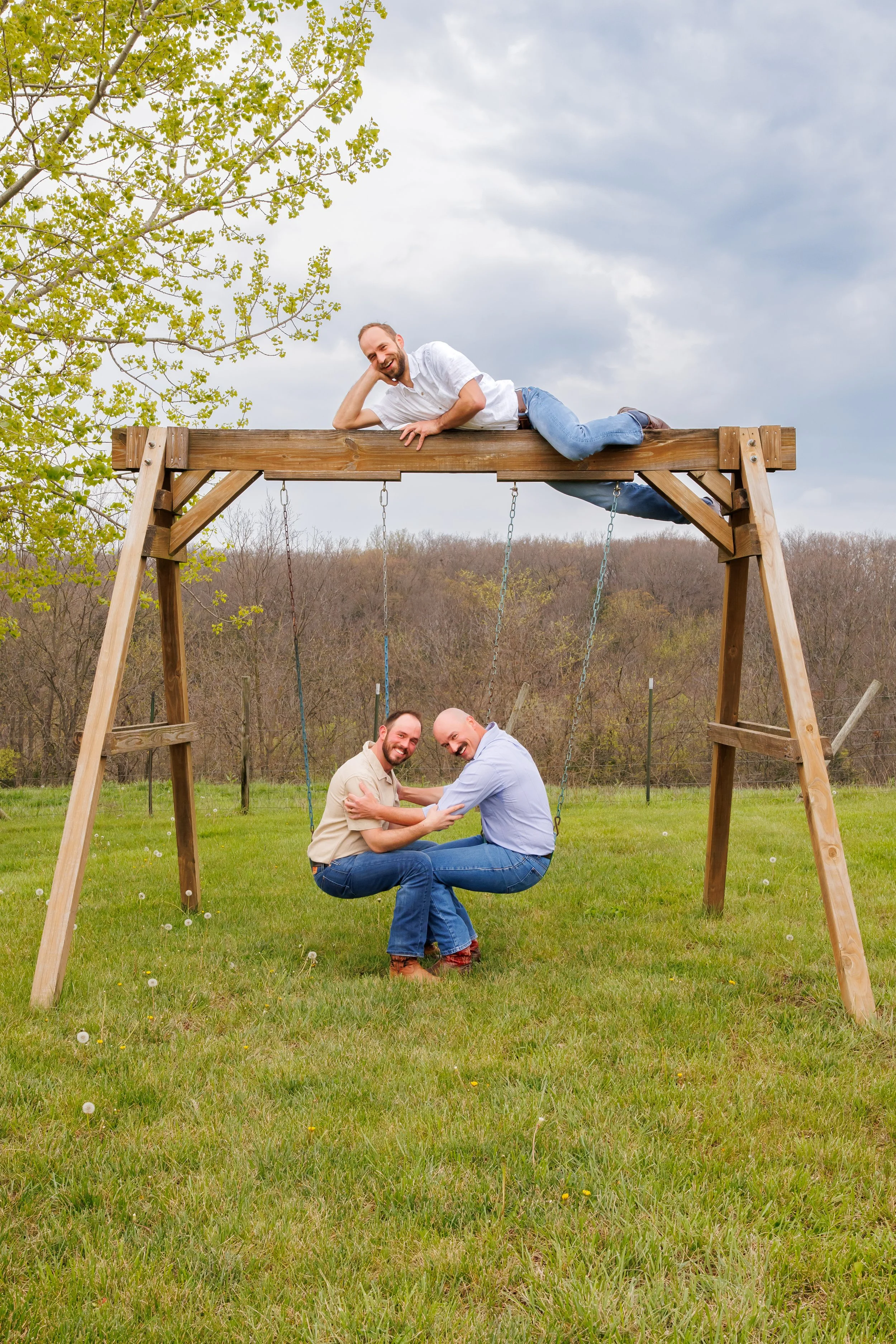 Two couples playing on a wooden swing set outdoors on a grassy field with trees and cloudy sky in the background. One man is lying on top of the swing set while the other couple is sitting on the swings.