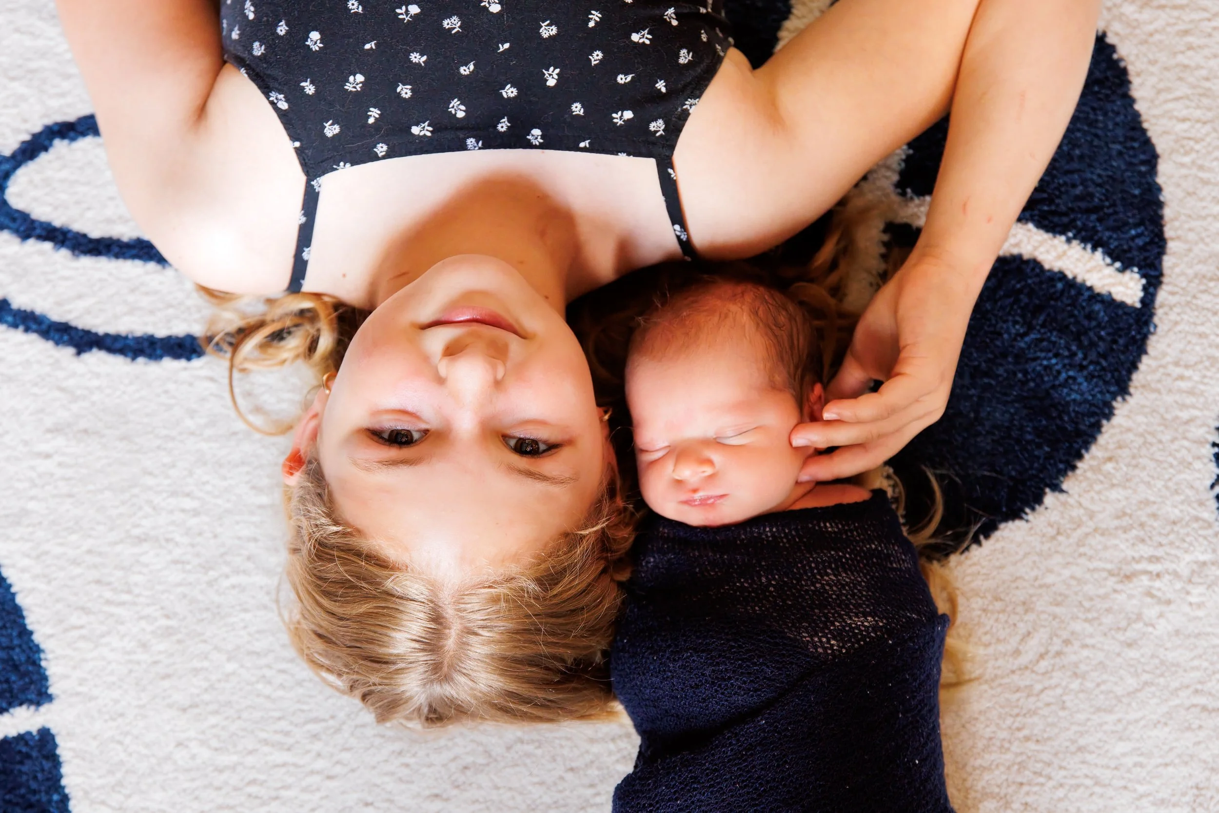 A woman with wavy blonde hair lying on the floor with a toddler, both on a white and blue striped rug. The woman is wearing a black dress with white flower patterns, and the toddler has curly hair and is resting with their eyes closed, with a hand touching their face.