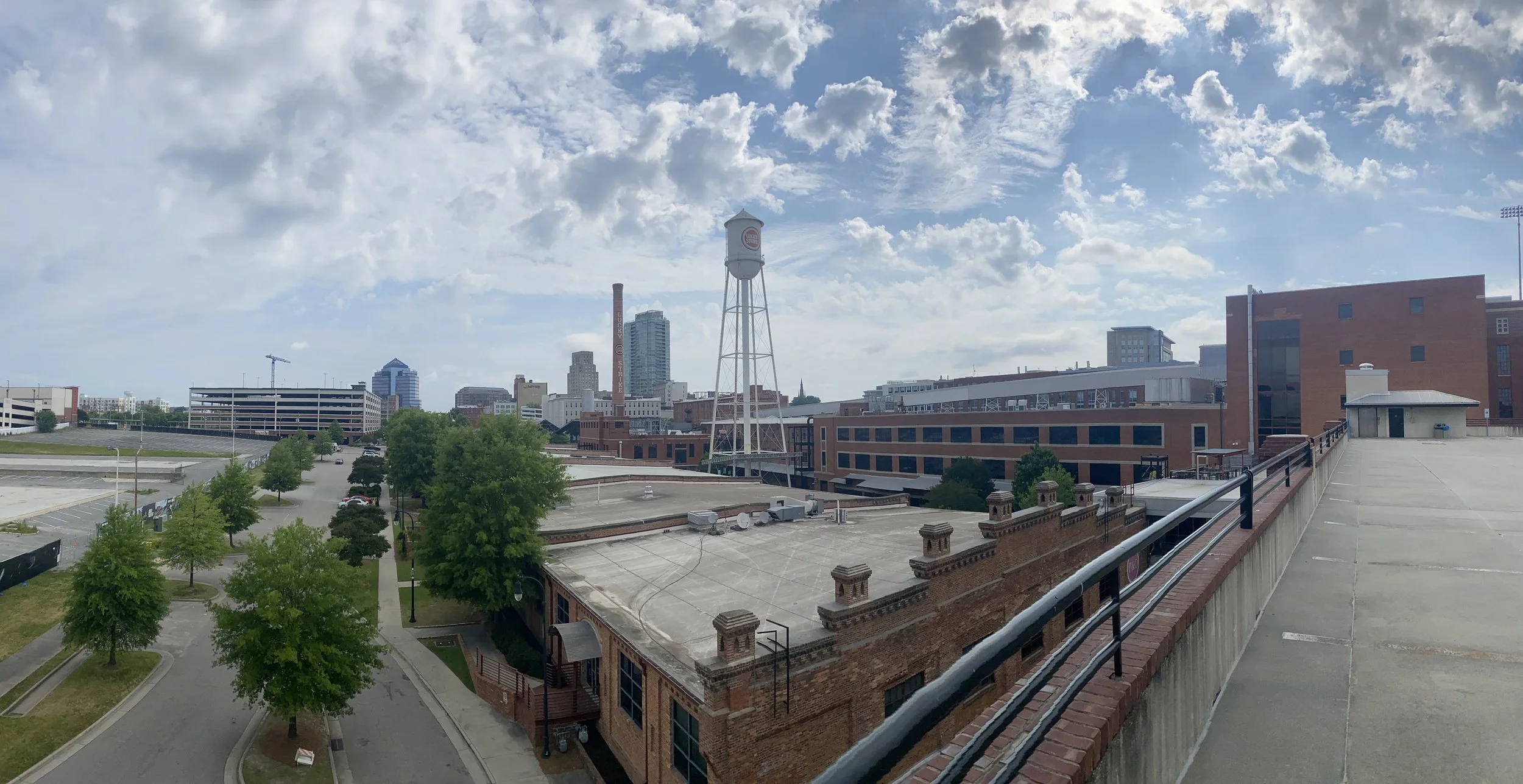Cityscape view from a rooftop with buildings, trees, parking lot, and partly cloudy sky.