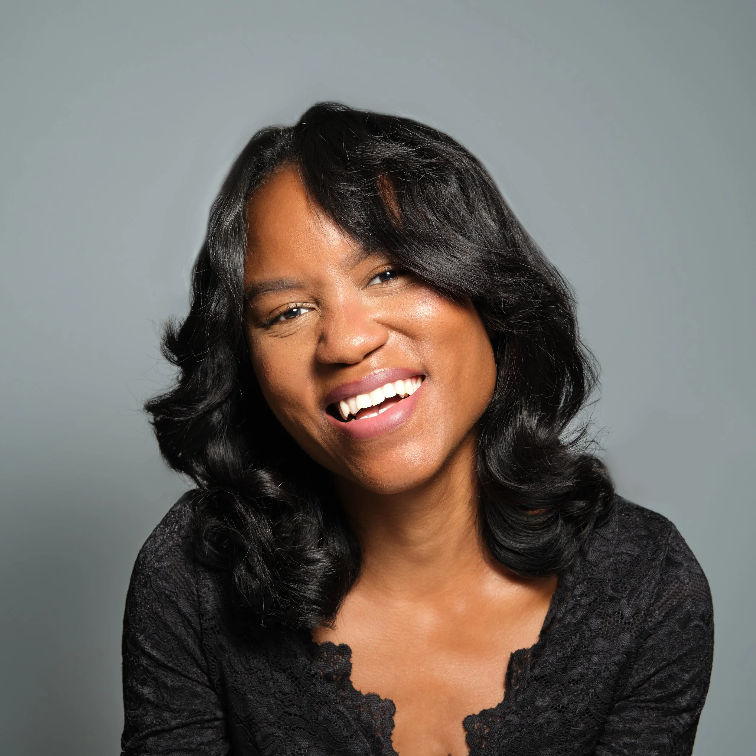 A woman with shoulder-length black curly hair smiling and wearing a black lace top against a gray background.