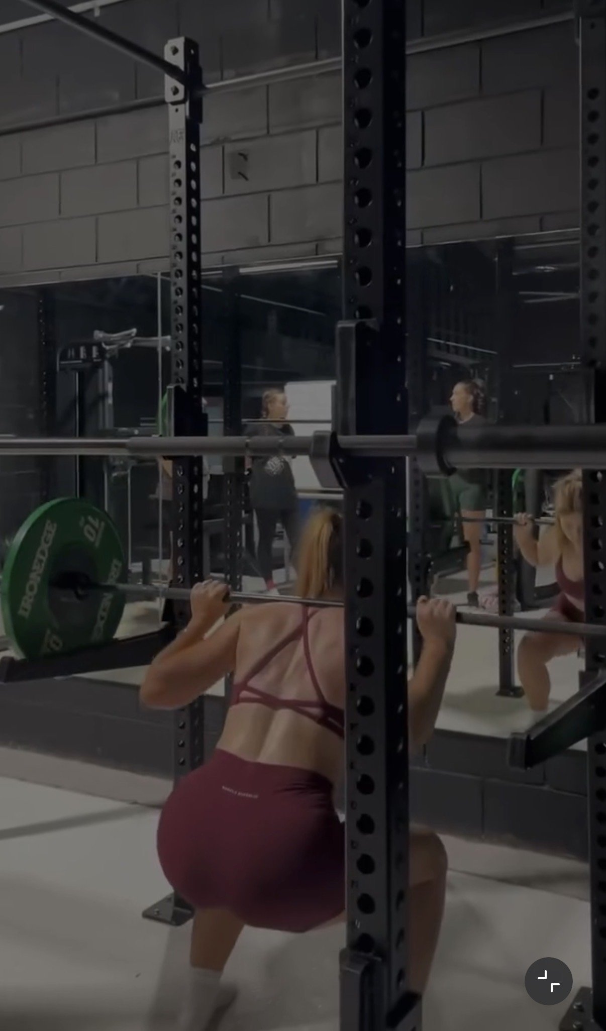 A woman wearing a sports bra and shorts performing a squat exercise in a gym with a barbell loaded with green weight plates.