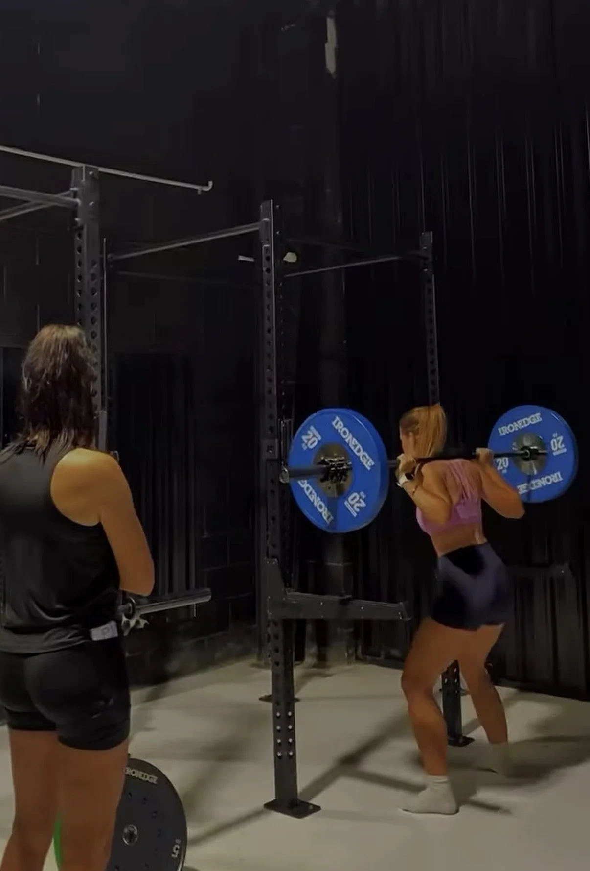 A woman is performing a squat with a barbell with blue plates at a gym. A person is observing her, and black gym equipment is visible in the background.