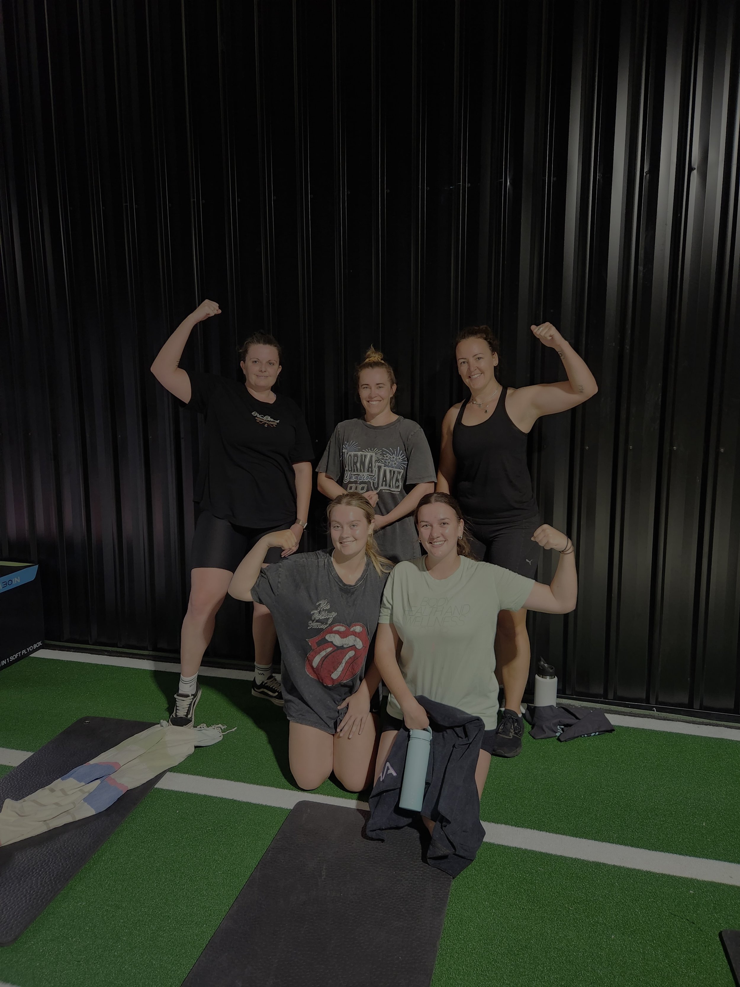 Group of six women celebrating fitness success in gym, lifting their arms in victory. Four women are standing, two are kneeling in front. The background features a black wall and green flooring.
