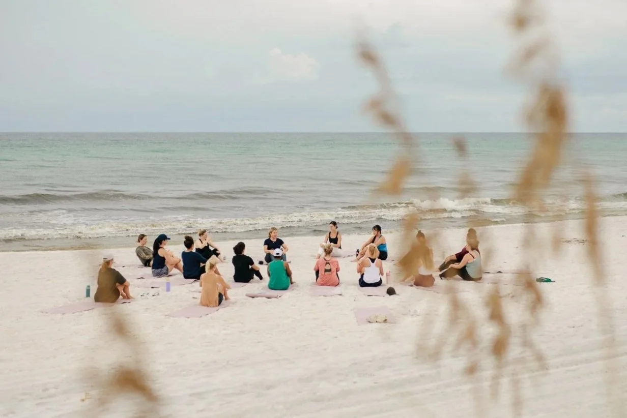 Assorted salad toppings and ingredients on a table, including potato chips, tortilla chips, two types of salsa, guacamole, chopped onions, chopped tomatoes, chopped red onions, a bowl of cooked grains, a bowl of dressed greens, a bowl of soup with beans, and a casserole dish with cooked beans.