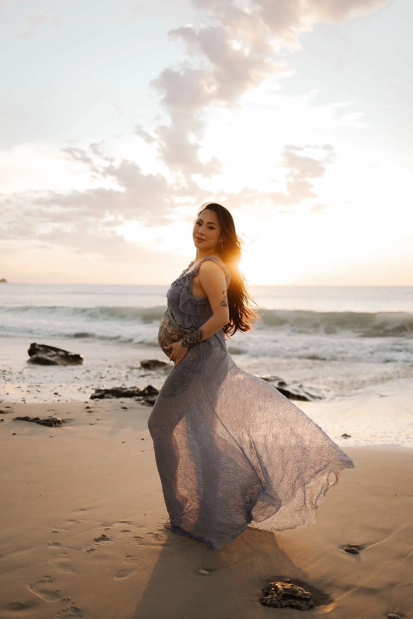 A pregnant Adelaide woman with tattoos on her arms and stomach, wearing a flowing dusty blue dress, stands barefoot on a sandy beach at Port Willunga during sunset, with the ocean and a partly cloudy sky in the background.