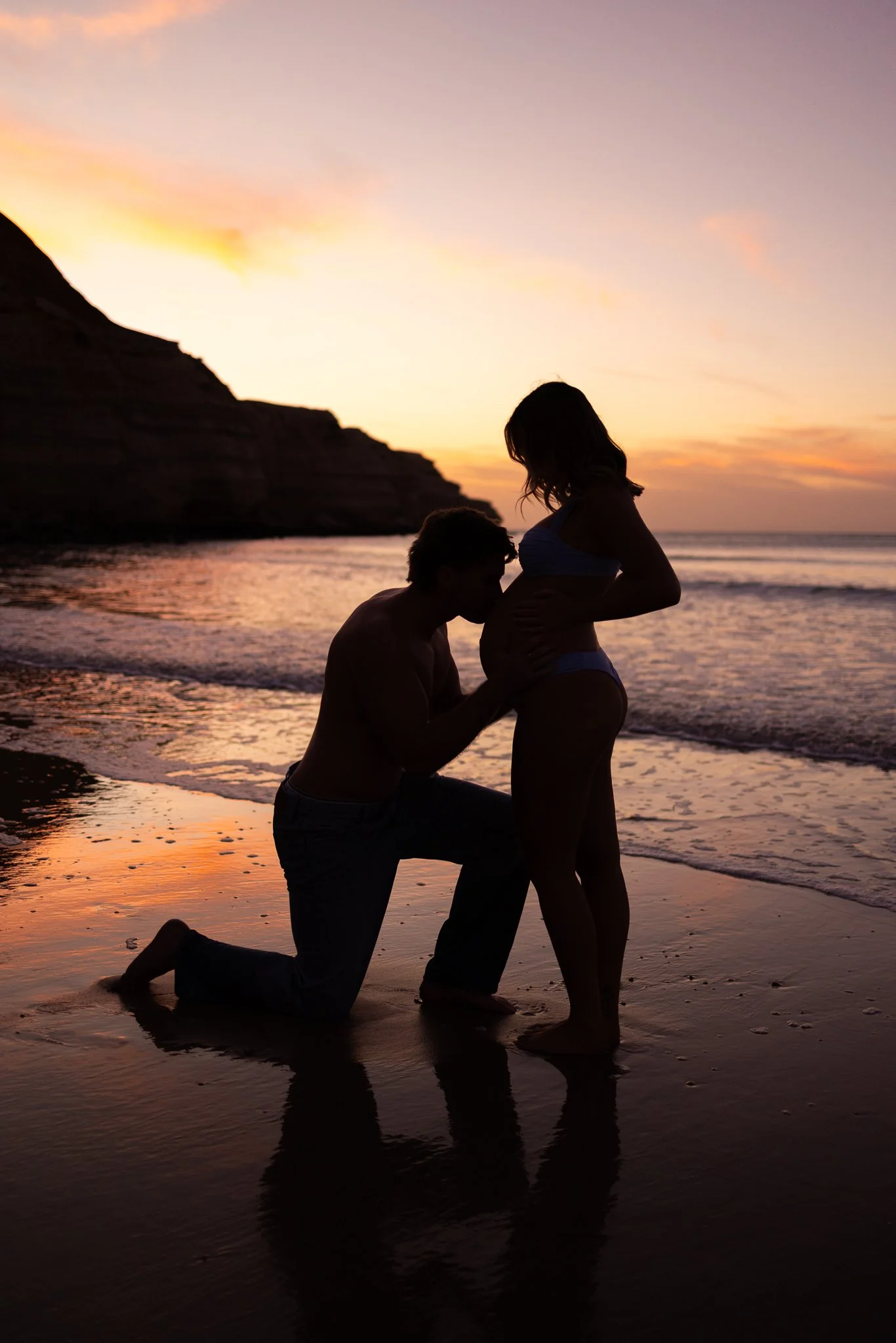 A dreamy silhouette of a man kissing a pregnant woman's baby bump on Adelaide beach at sunset, with waves and a rocky cliff in the background.