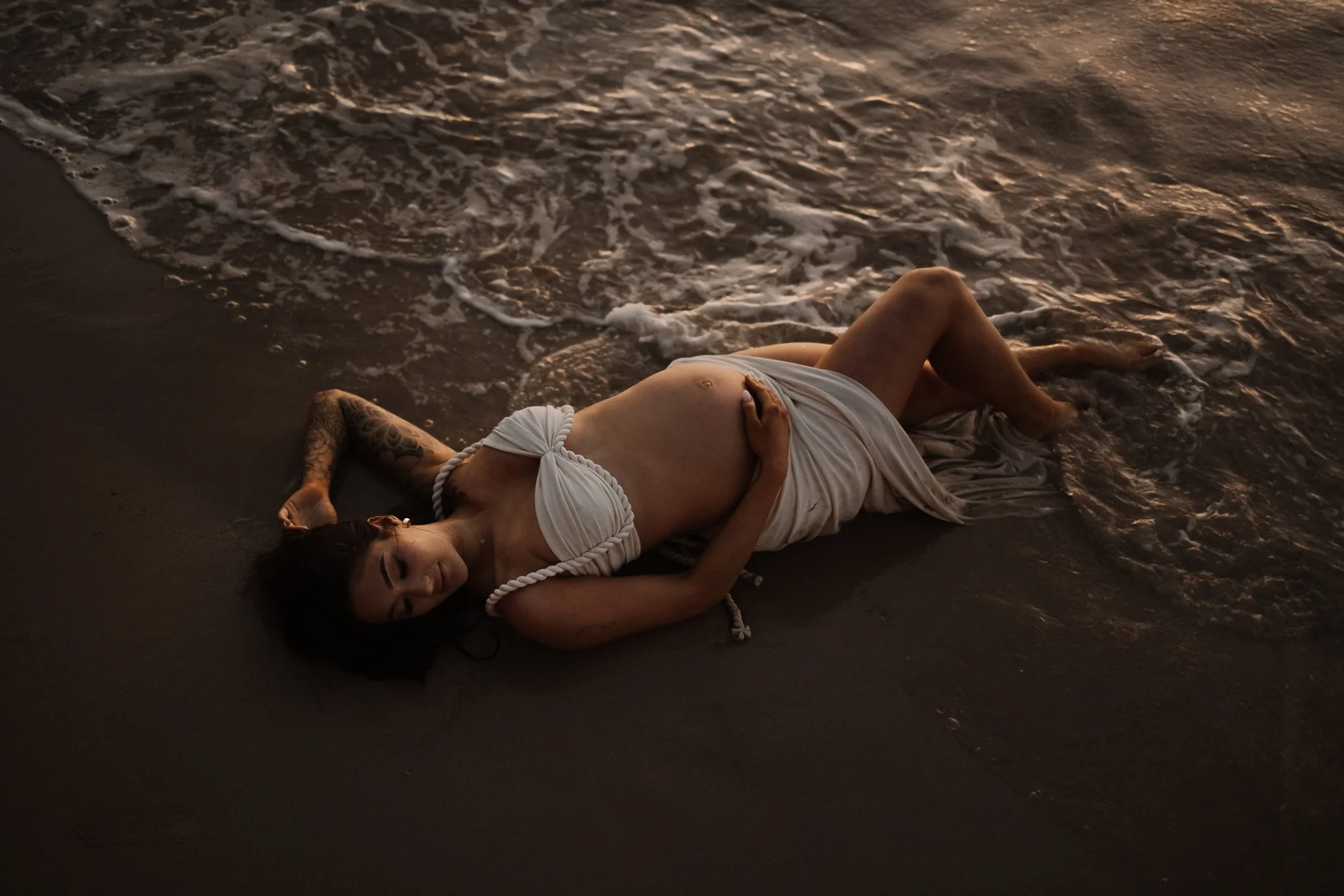 A pregnant woman lies on the beach at sunset, with her head turned to the side and her hand resting on her belly, wearing a white two piece top and skirt and surrounded by gentle waves for her maternity photoshoot.