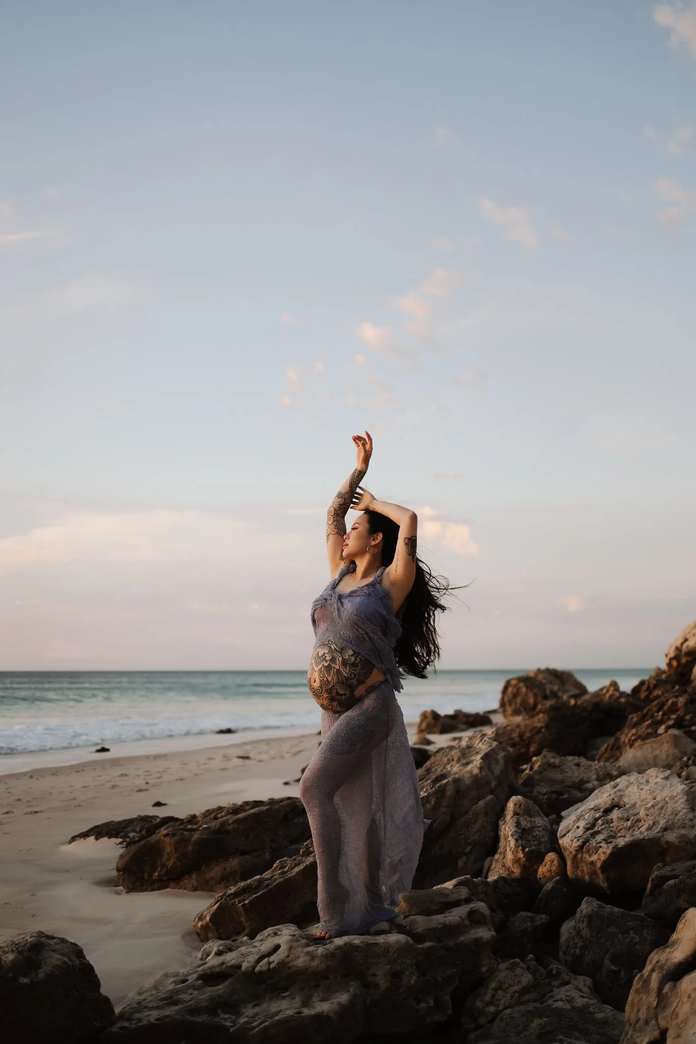 An Adelaide pregnant woman dressed in gray and lace clothing standing on rocks at the beach, with her hands raised above her head and looking to the side, during sunset.