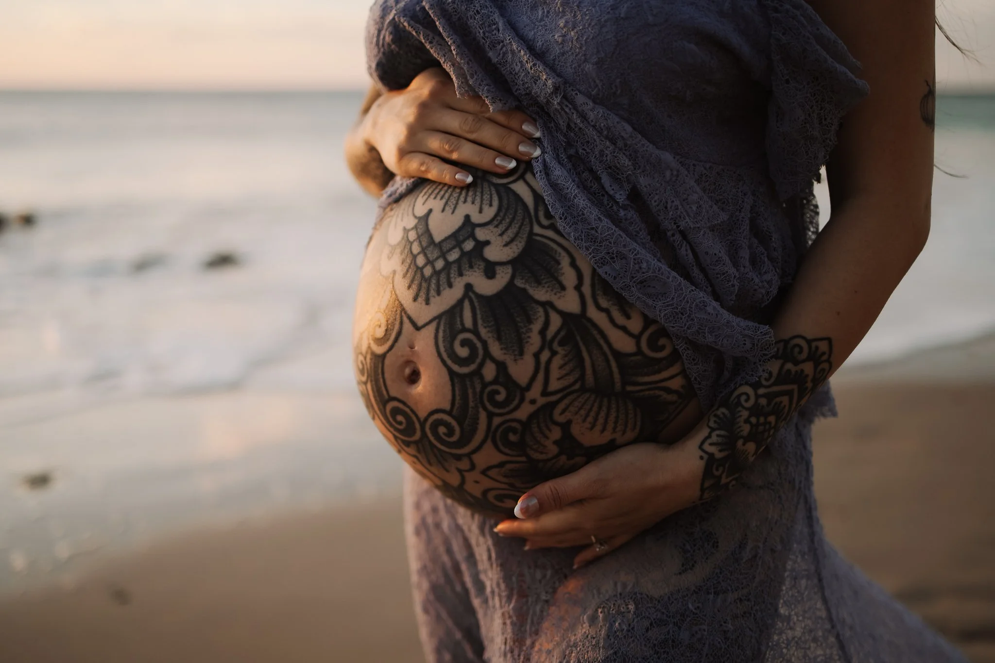 A pregnant belly photo of a woman with intricate tattoos on her belly and arm is holding her pregnant belly with both hands at the beach during sunset, with the ocean and sand visible in the background.