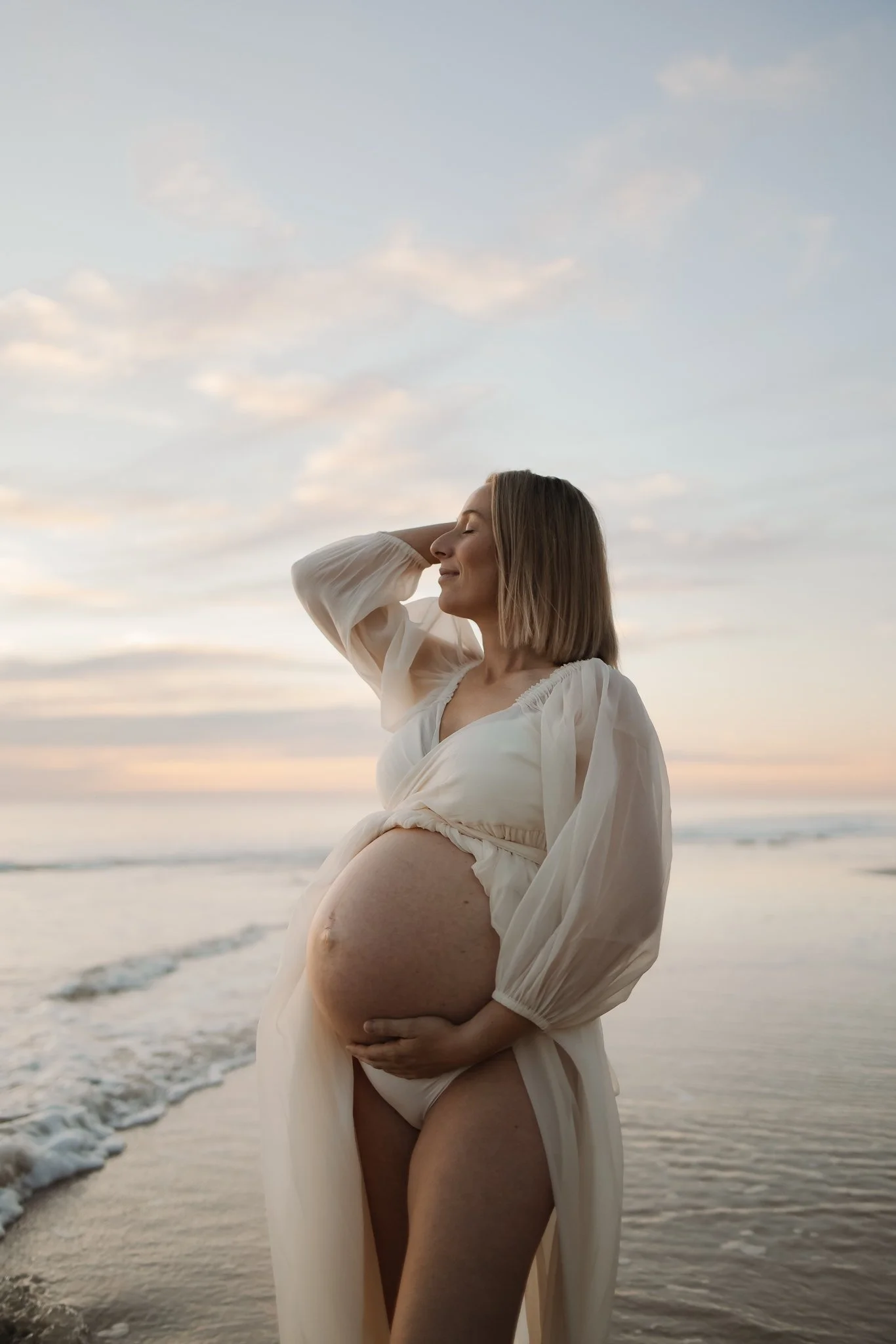 Pregnant woman in a flowing dress standing on the beach with her hand on her belly, enjoying the sunset with a calm ocean on an Adelaide beach in the background while having her maternity photos.