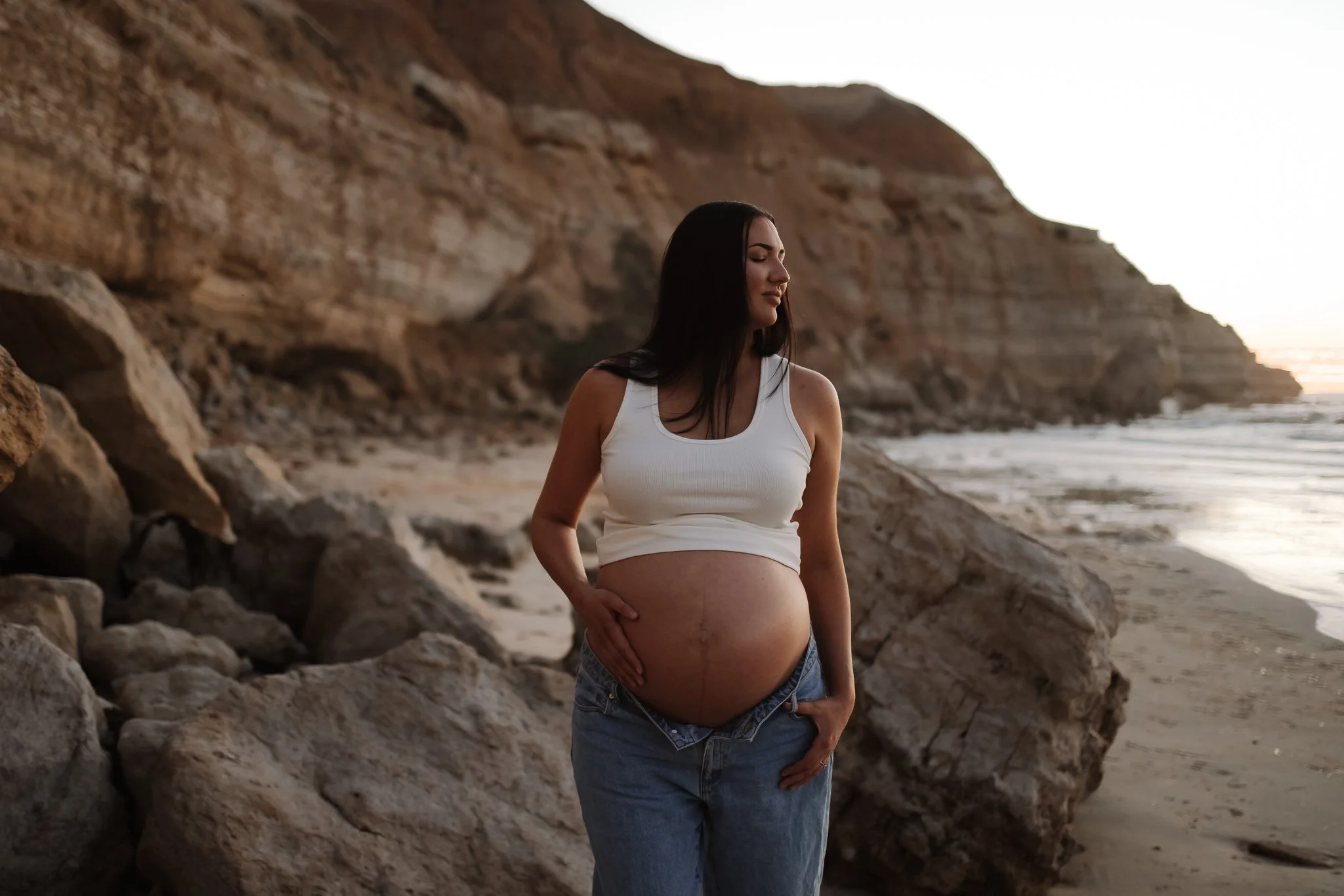women wearing a cropped tank and unbuttoned jeans with her pregnant belly exposed as she stands surround by the cliffs of Maslin Beach. Pregnant Women has a soft smile on her face as she closes her eyes soaking in her pregnancy.