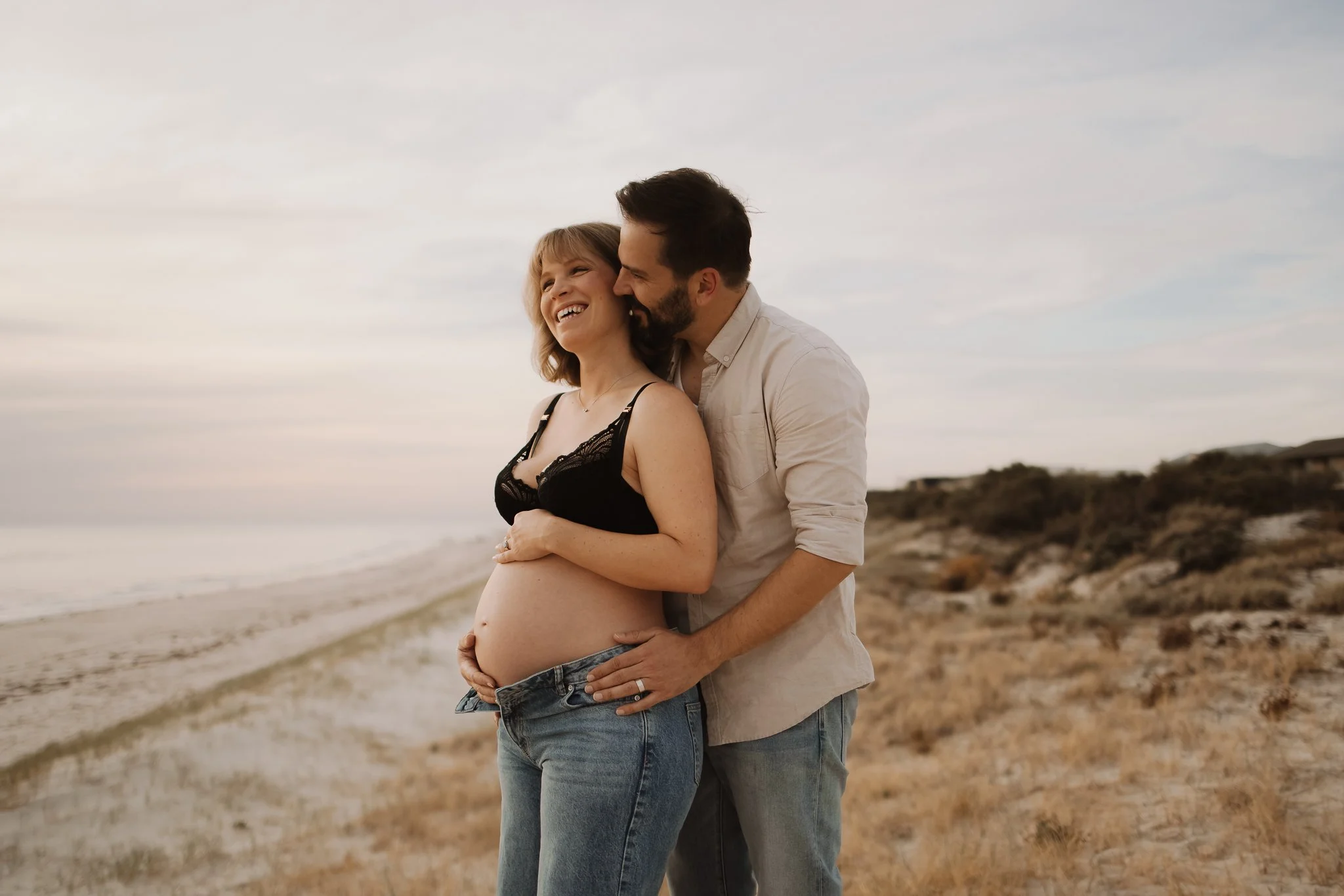 A pregnant woman and her partner standing on an Adelaide beach, Tennyson, smiling, with their hands on her pregnant belly, nature and ocean in the background during sunset at their maternity photoshoot.