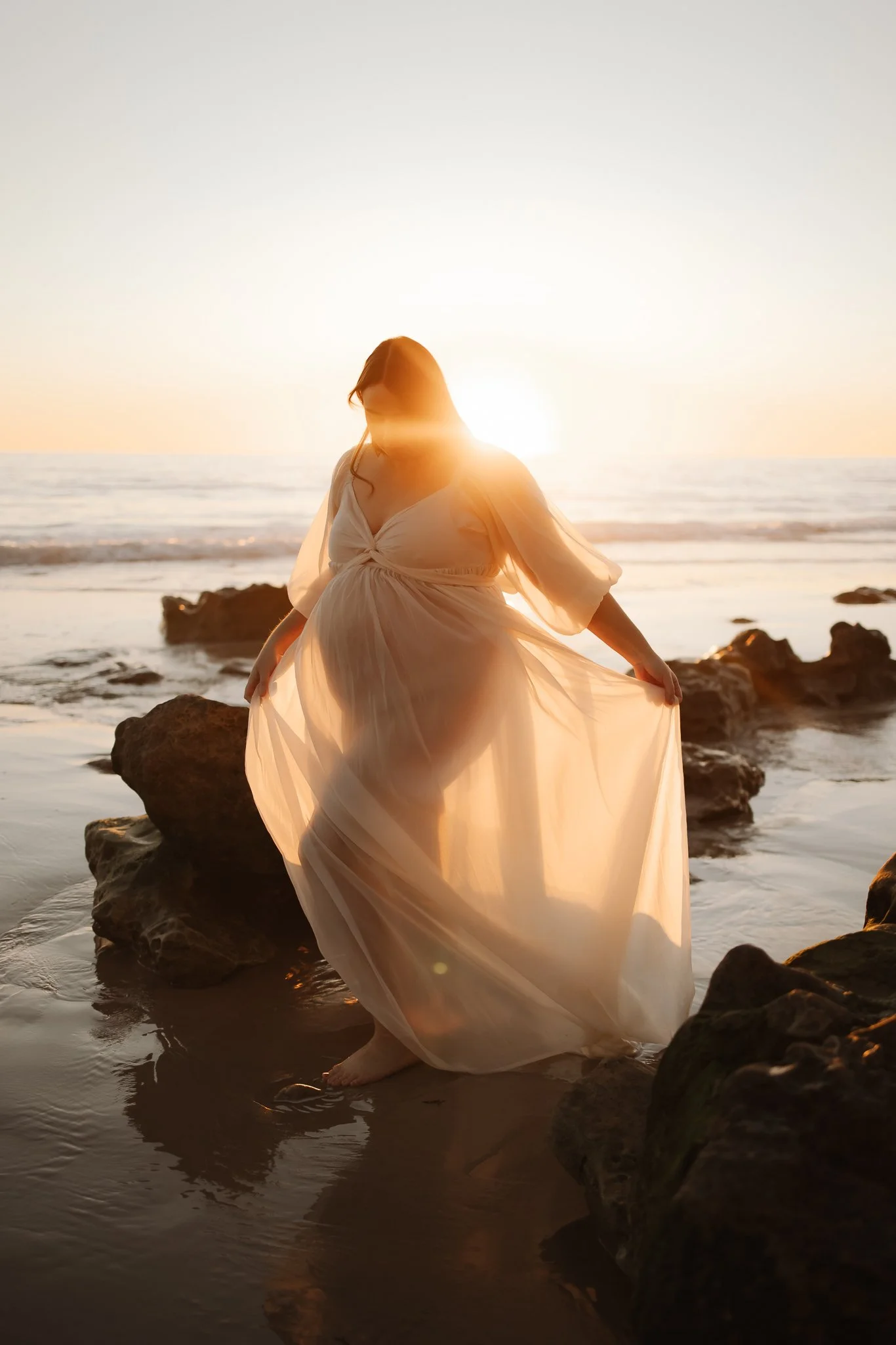 A pregnant woman in a flowing white sheer dress stands on rocks at the beach during sunset, with the sun glowing golden behind her at her Maternity photoshoot at a beach south of Adelaide.