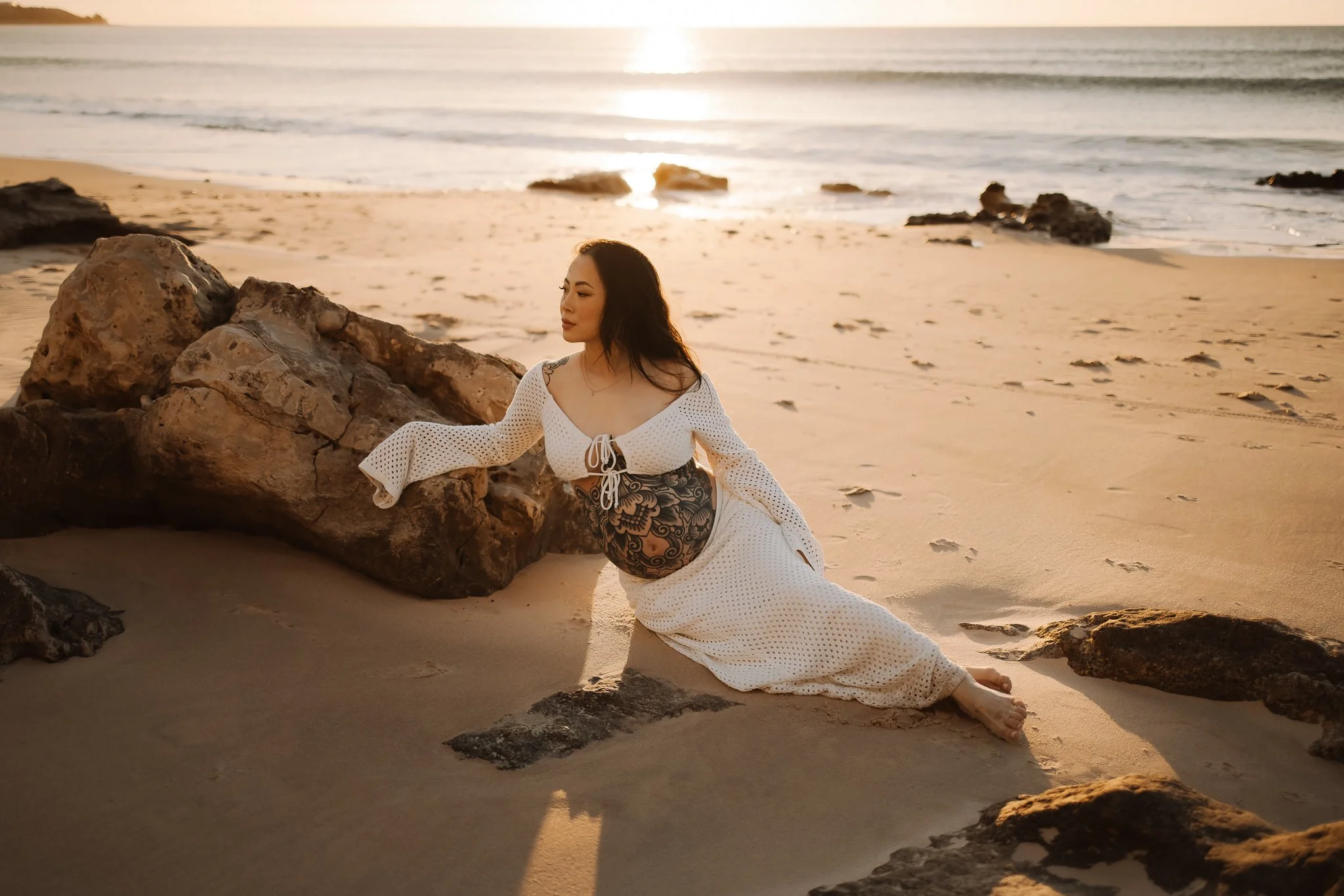 Photo of a pregnant Adelaide woman with long dark hair sitting on the sand near rocks at an Adelaide beach during sunset, wearing a white dress and showing her tattooed belly.