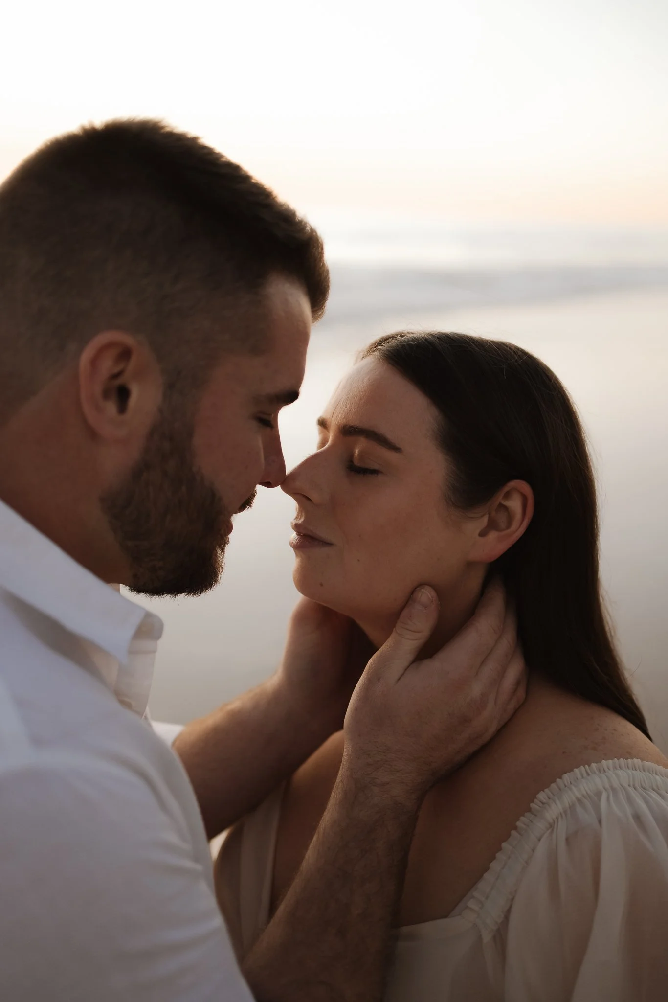 A pregnant couple sharing a tender, intimate moment on an Adelaide beach at sunset, with their noses touching and eyes closed, expressions of affection.