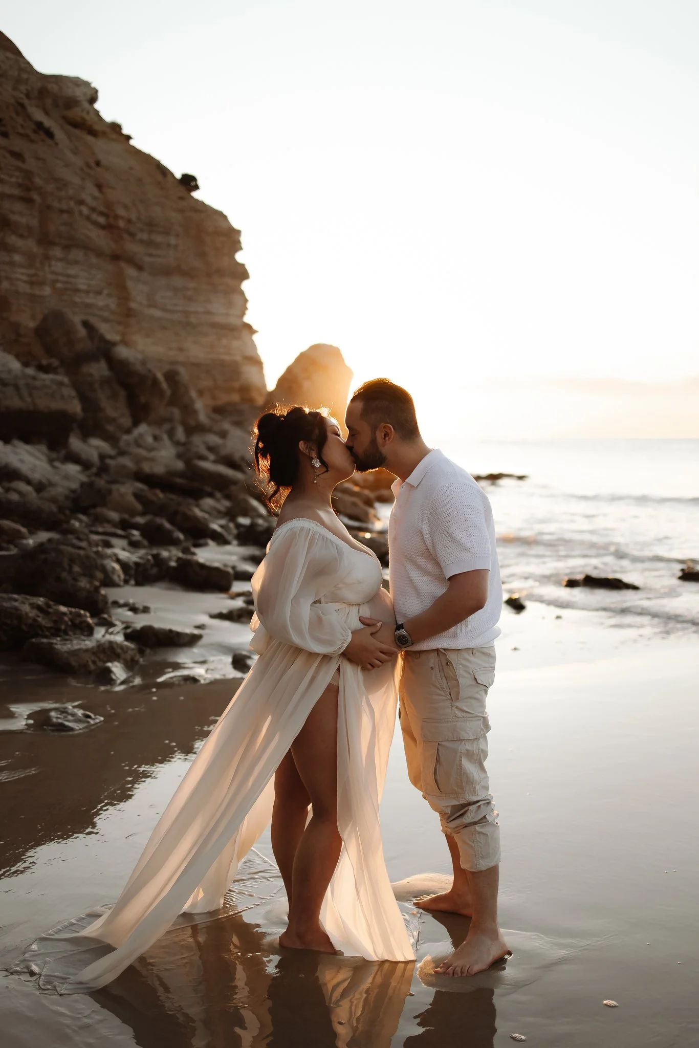 pregnant women in sheer flowing dress and husband kiss in front of the cliffs at Maslin Beach, South Australia.