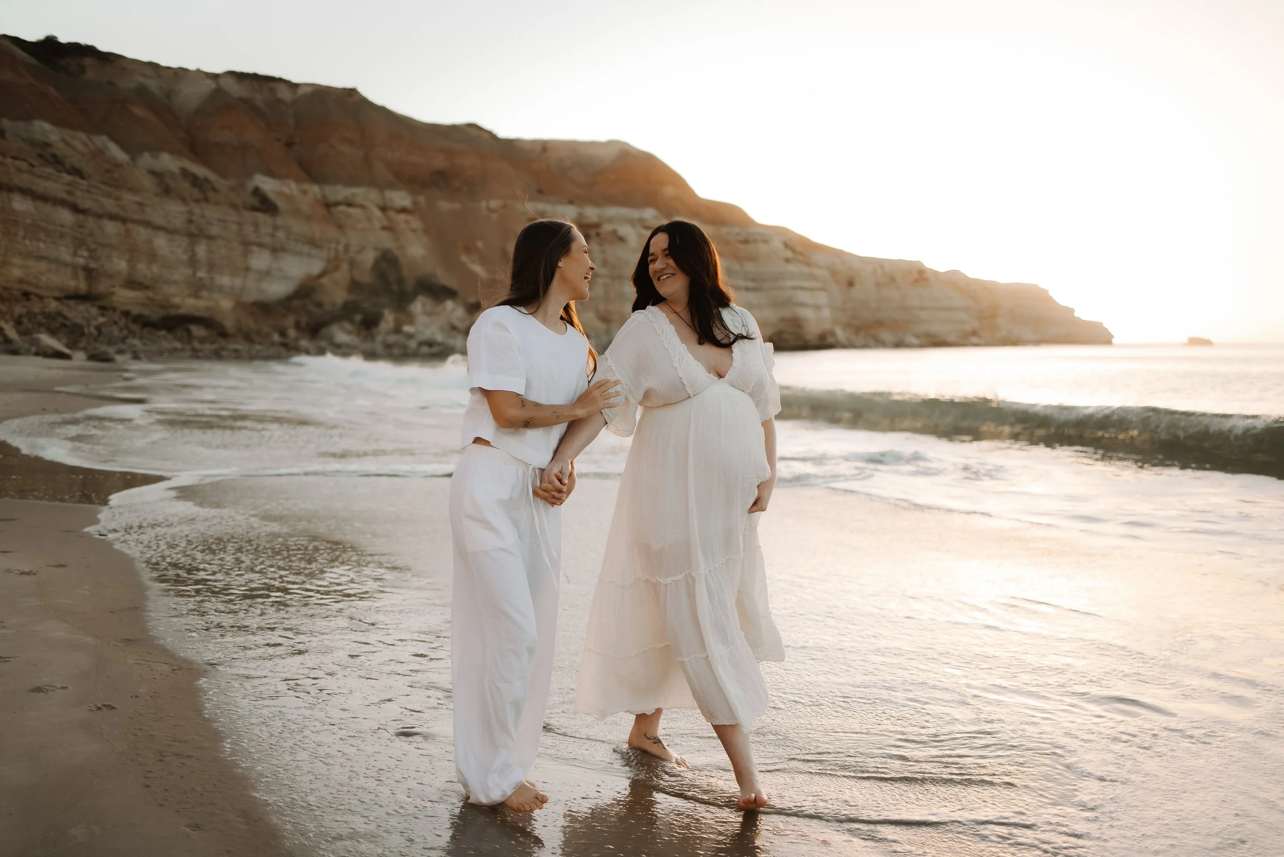 two married women walk on an Adelaide  beach embracing each other at sunset. One women is pregnant their first child.